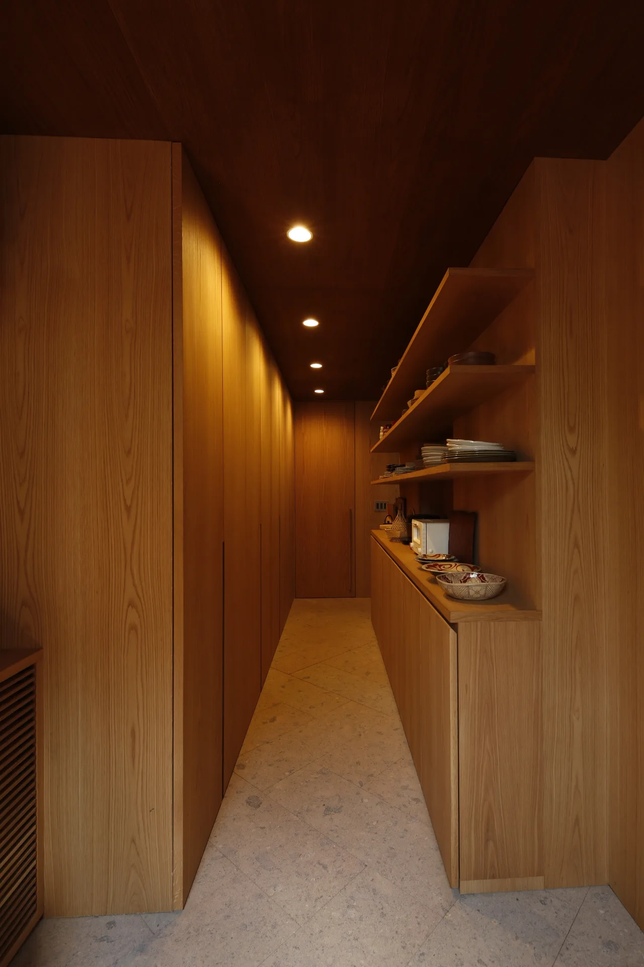 A narrow wood-paneled kitchen corridor features built-in cabinets, open wooden shelves holding ceramic dishware, and a stone tiled floor.
