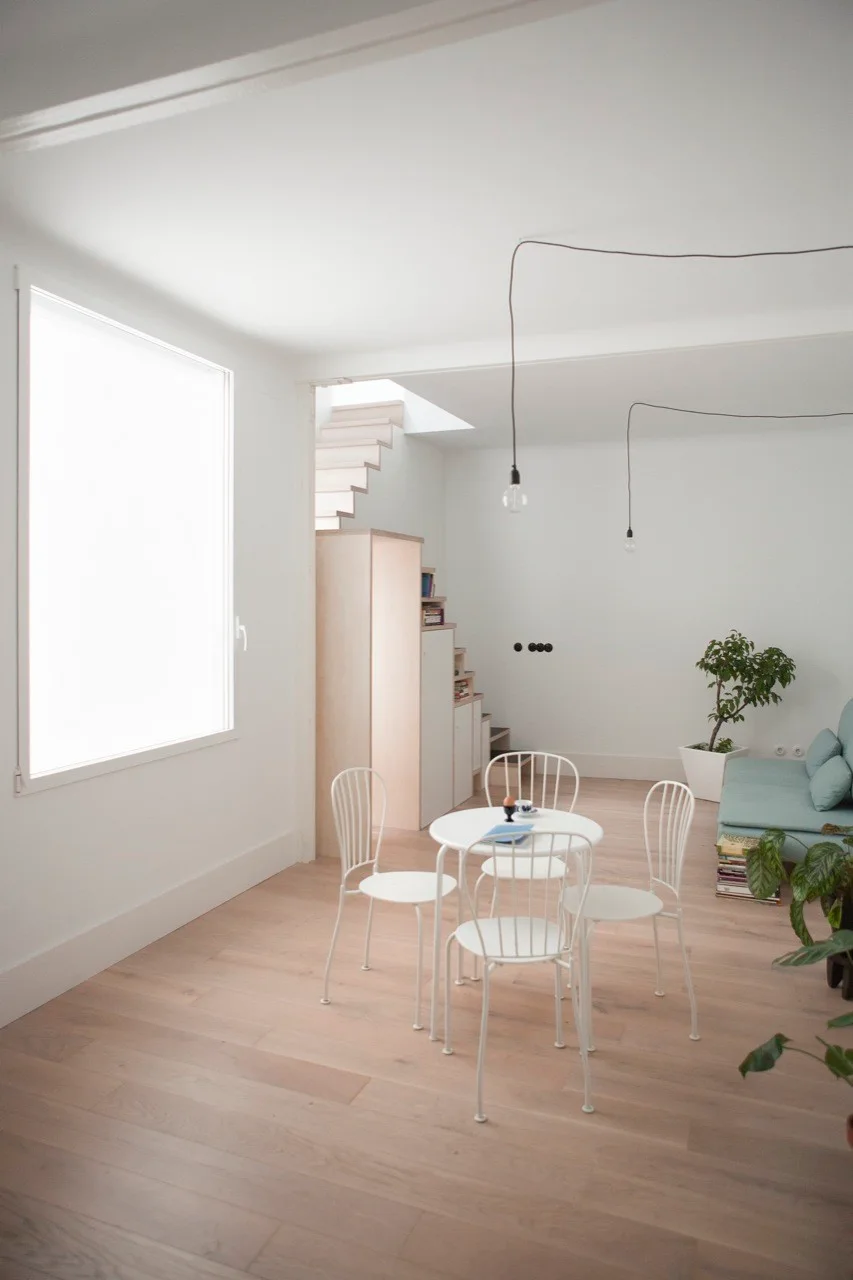 A bright open-plan living and dining room features a white metal round table with chairs, light wood floors, and a light blue sofa in the background.