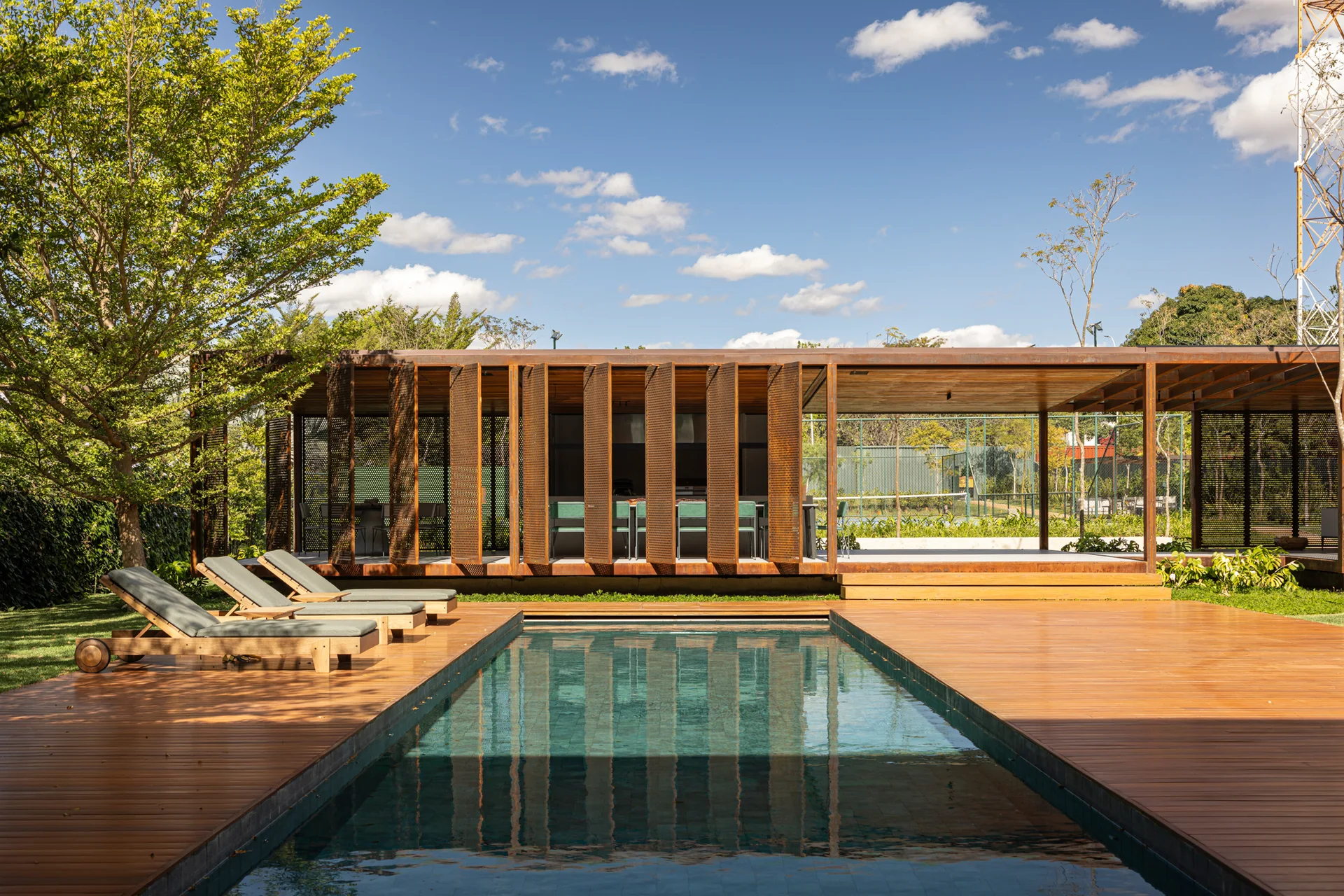 A rectangular swimming pool surrounded by a wood deck and an outdoor pavilion with rust-colored perforated metal panels.