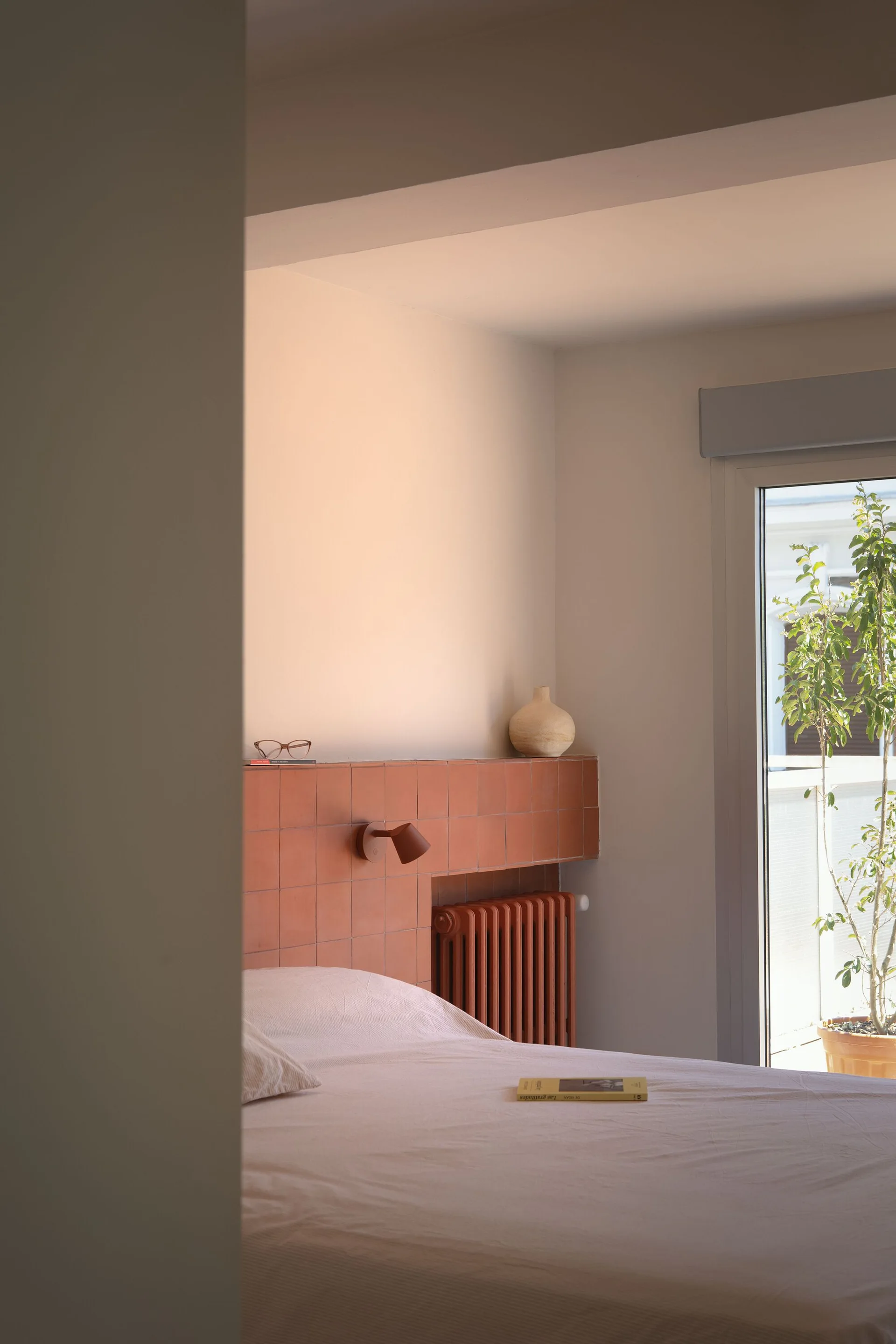 A minimalist bedroom features a bed with white sheets next to a terracotta tiled headboard ledge, a matching radiator, and a glass balcony door.