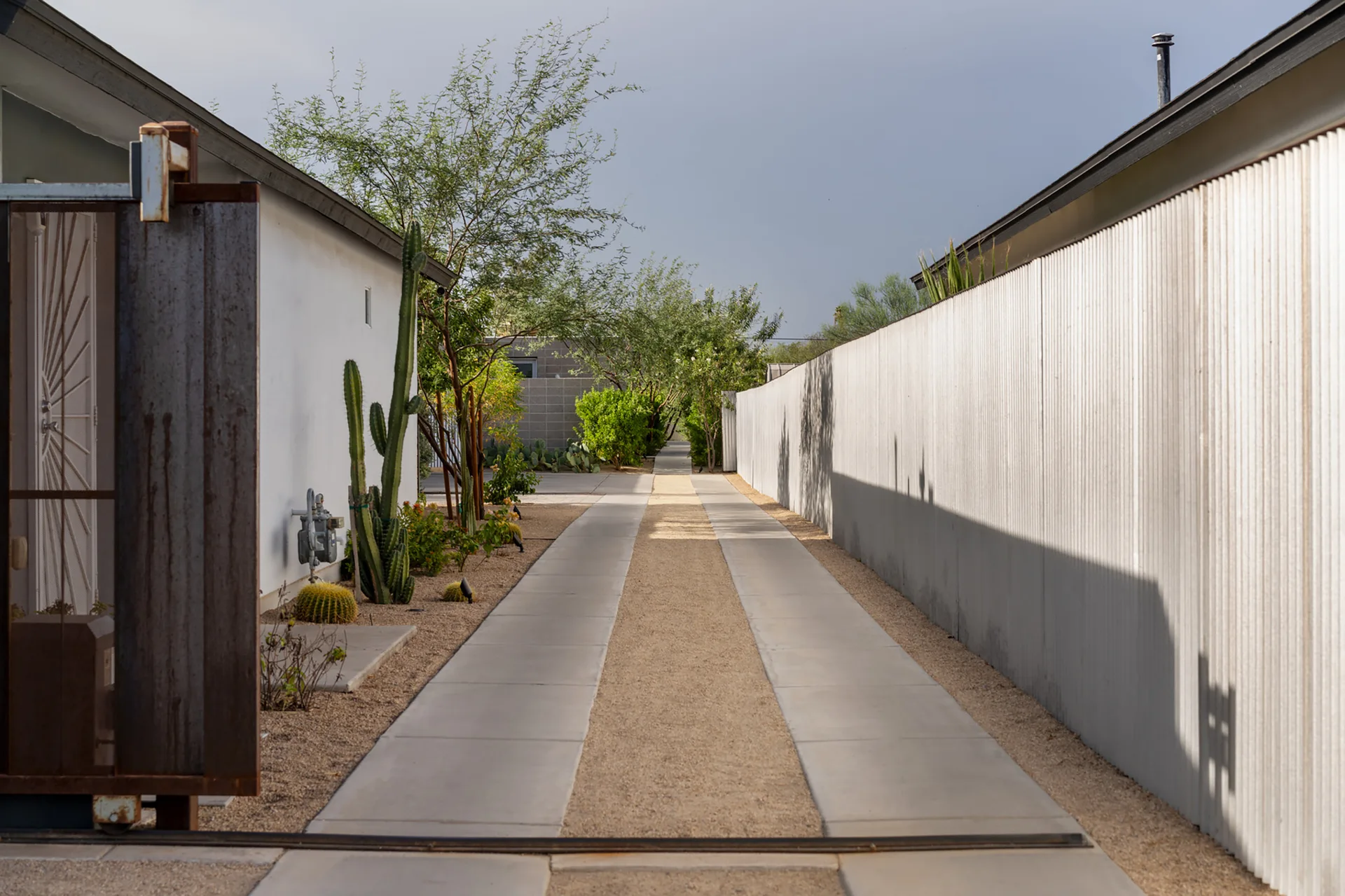 A concrete strip driveway runs between a white building with cacti and a long corrugated metal fence under a clear sky.
