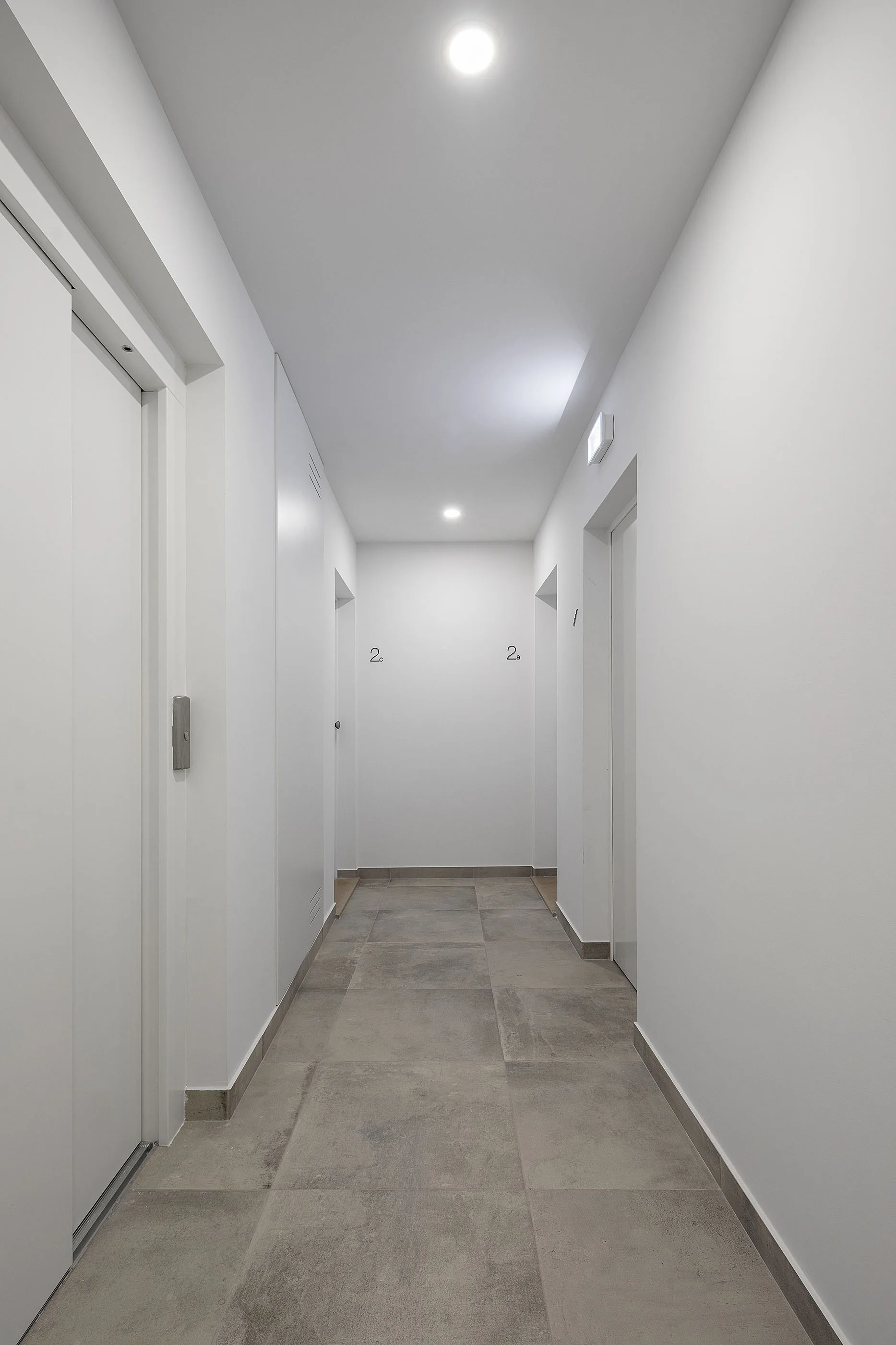 A long minimalist hallway features bright white painted walls, matching flush-mount white doors, recessed ceiling lights, and large grey floor tiles.