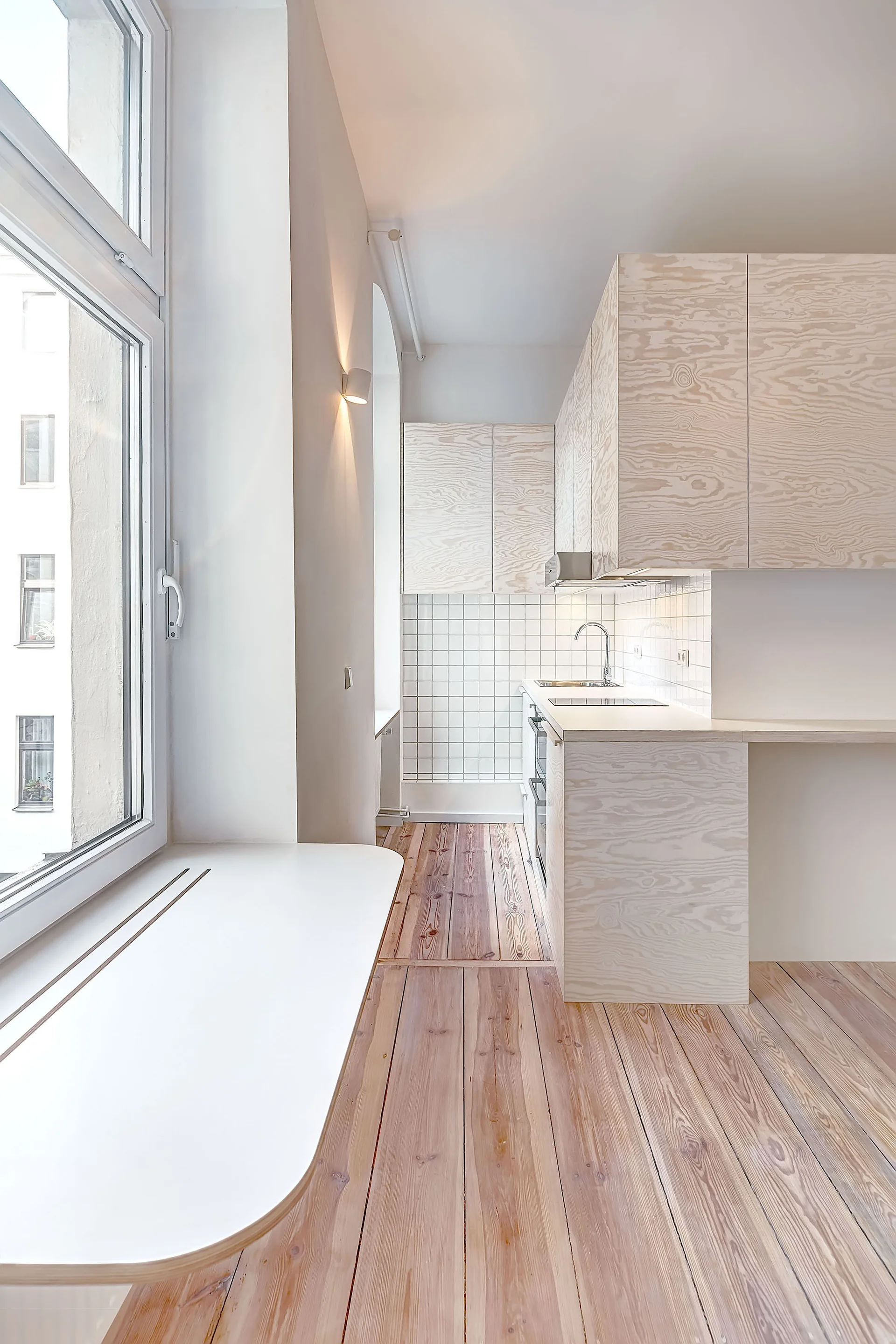A minimalist compact kitchen features light plywood cabinets, a white square grid tile backsplash, and wide-plank wood flooring.