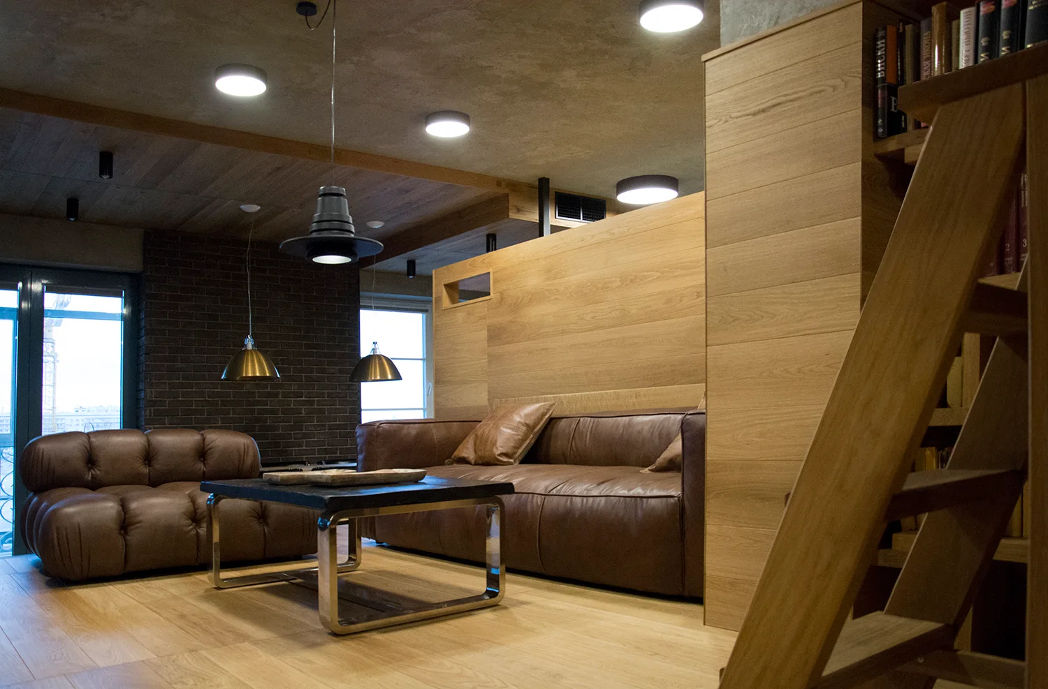 A modern industrial living room featuring a brown leather sofa, matching armchair, horizontal wood paneling, and a dark brick wall.