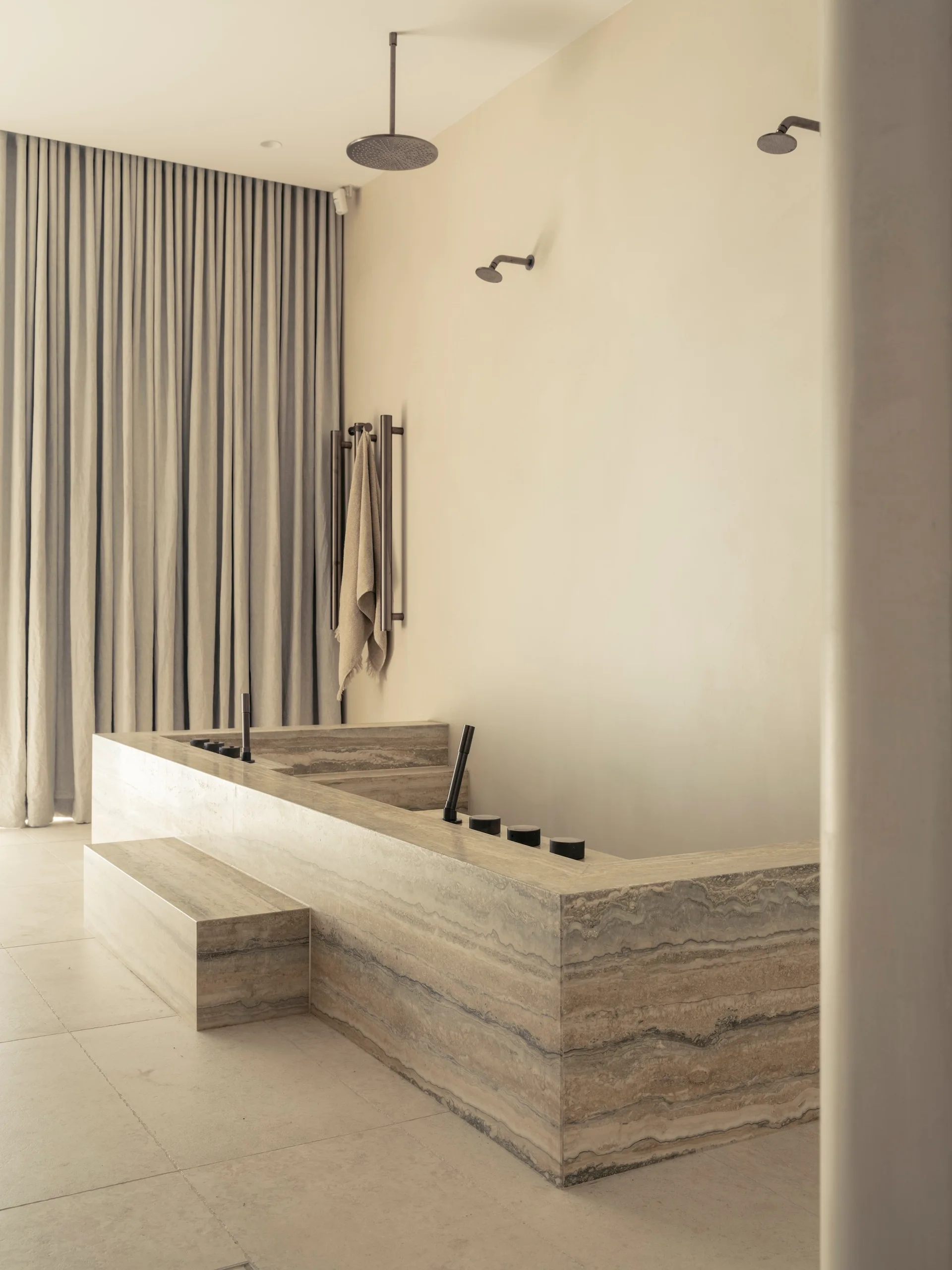 A minimalist bathroom features a large travertine stone bathtub with steps, matte plaster walls, and ceiling-mounted shower heads.