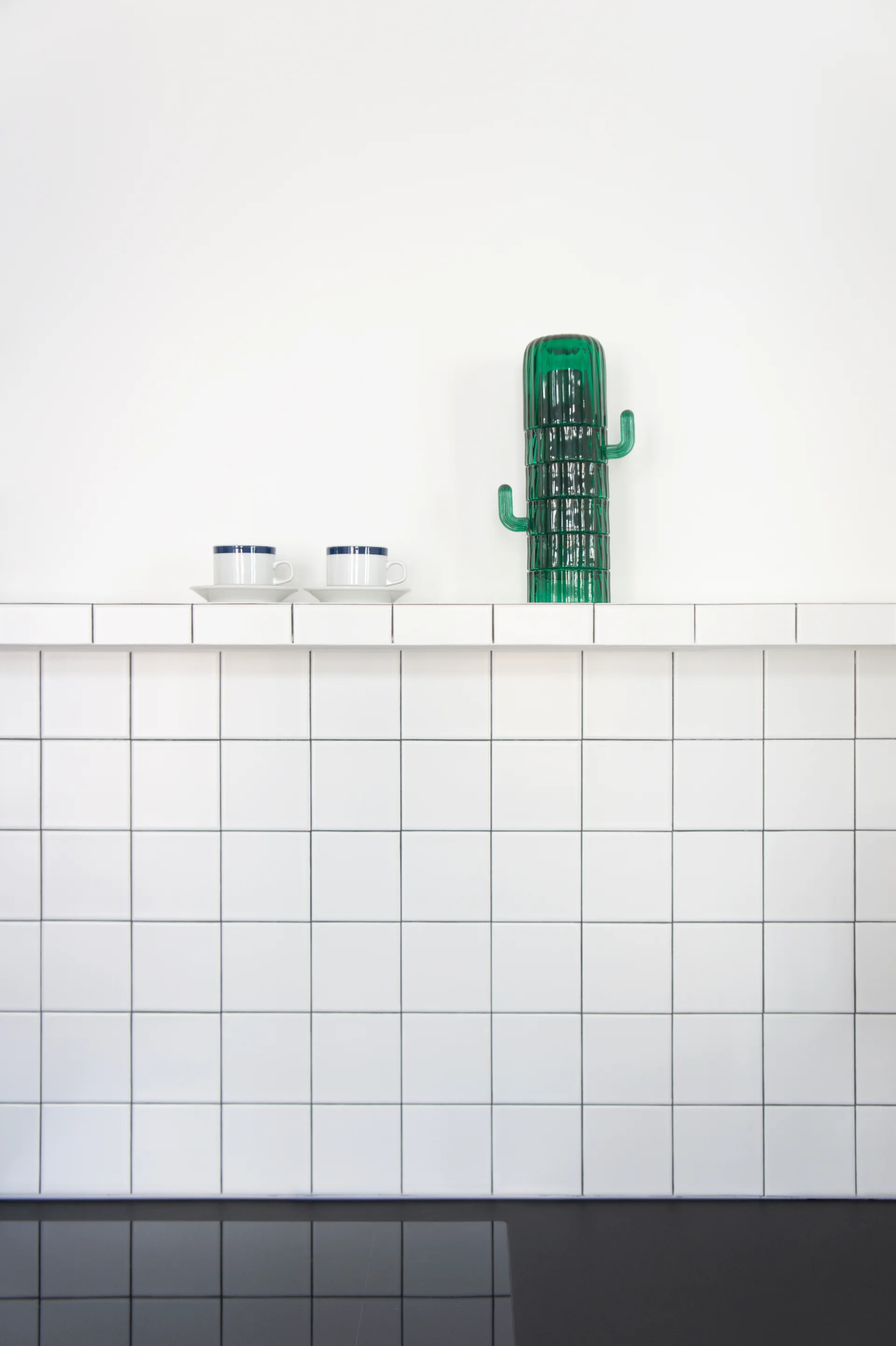 A white tiled kitchen backsplash features an integrated tiled shelf holding two cups and a green glass cactus sculpture.