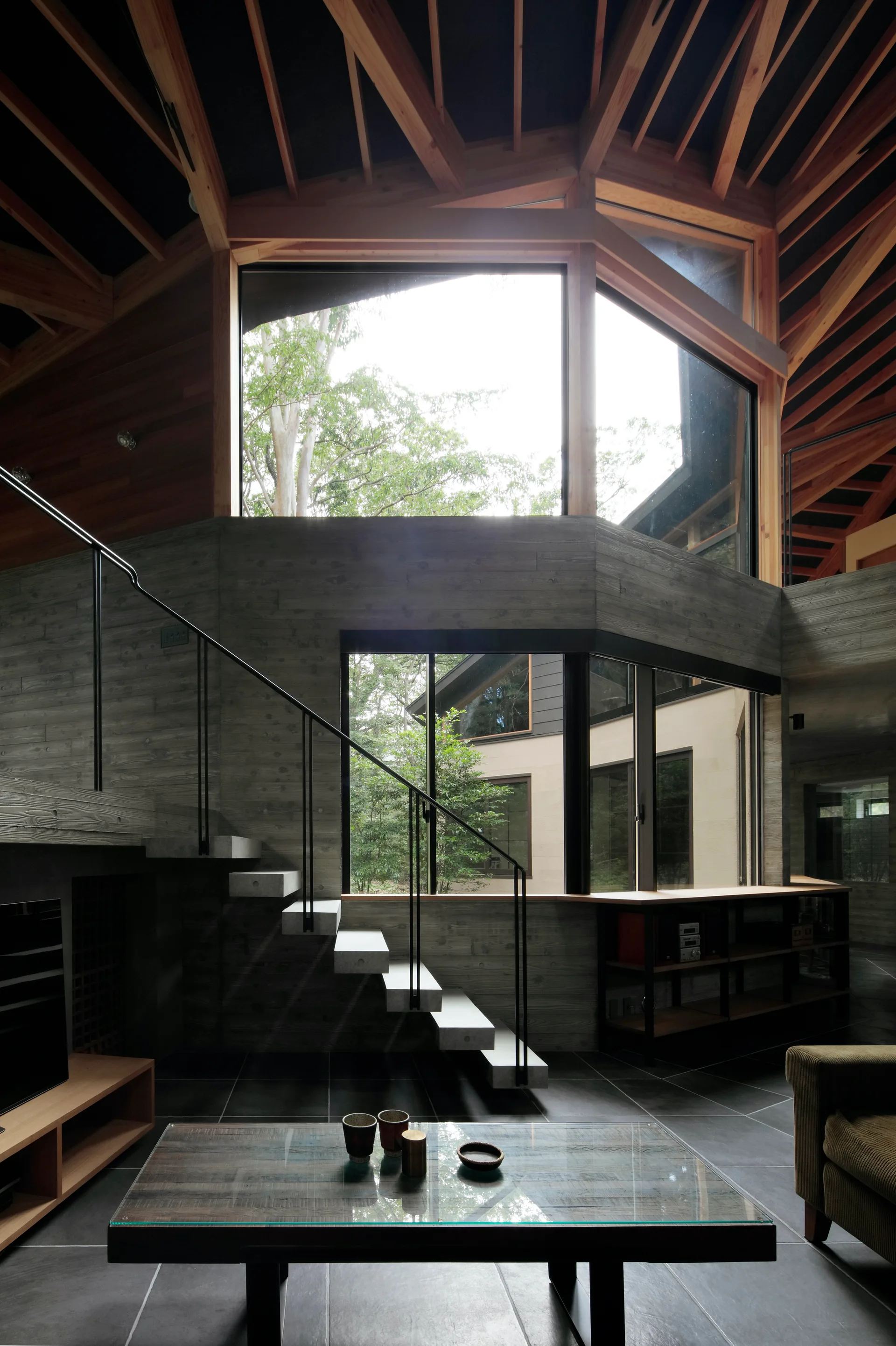 A modernist living space featuring board-formed concrete walls, a floating concrete staircase with a black metal handrail, and exposed wood ceiling rafters.