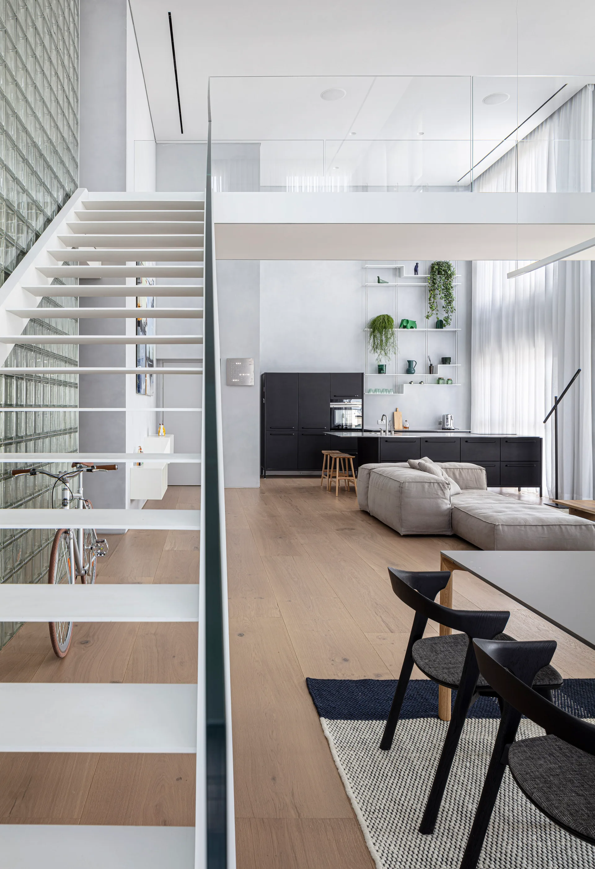 A modern minimalist loft features a white open-tread staircase against a translucent glass block wall, overlooking an open living room.