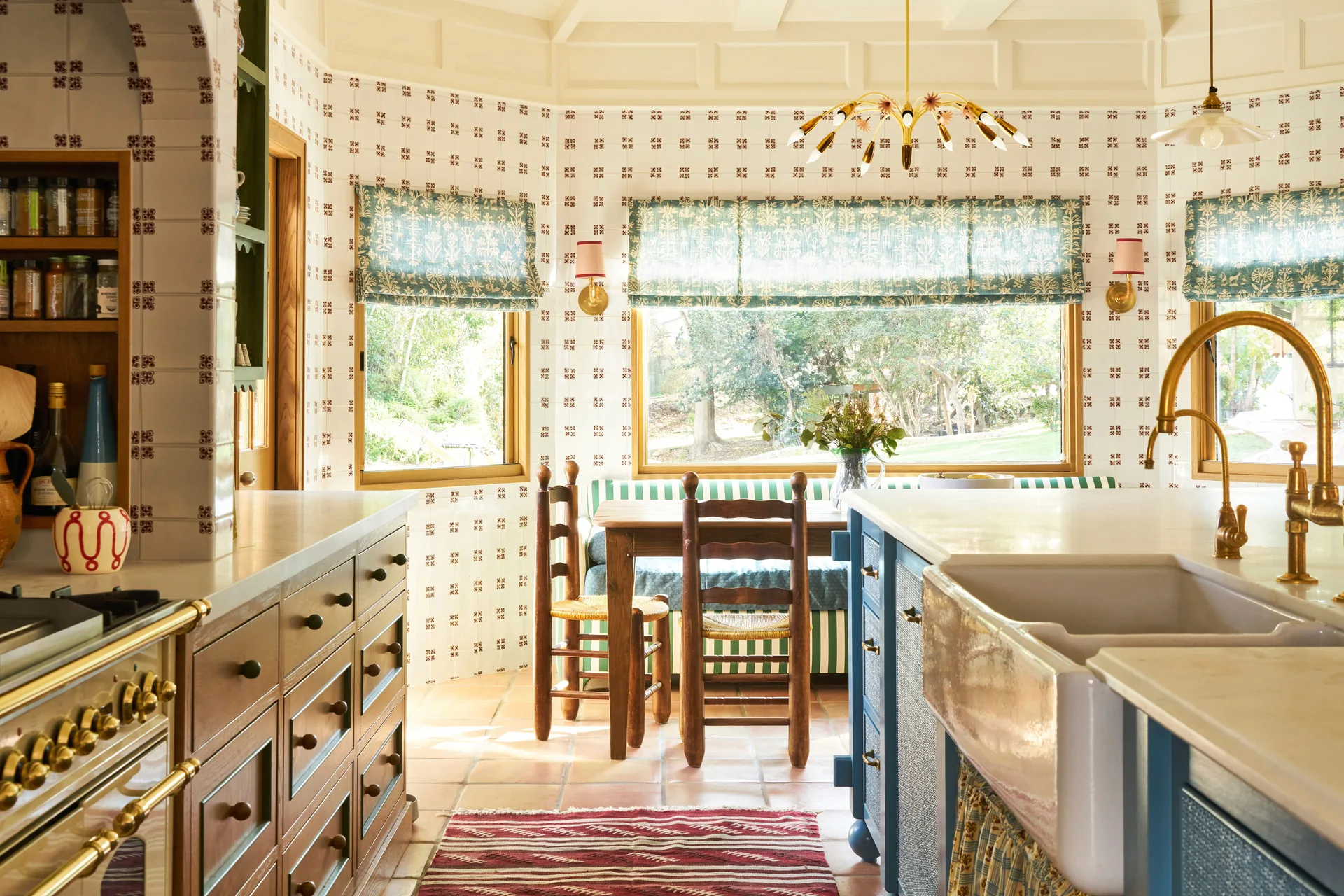 A sunlit kitchen features a breakfast nook with wooden furniture, patterned wall tiles, terracotta flooring, and a blue island with a farmhouse sink.