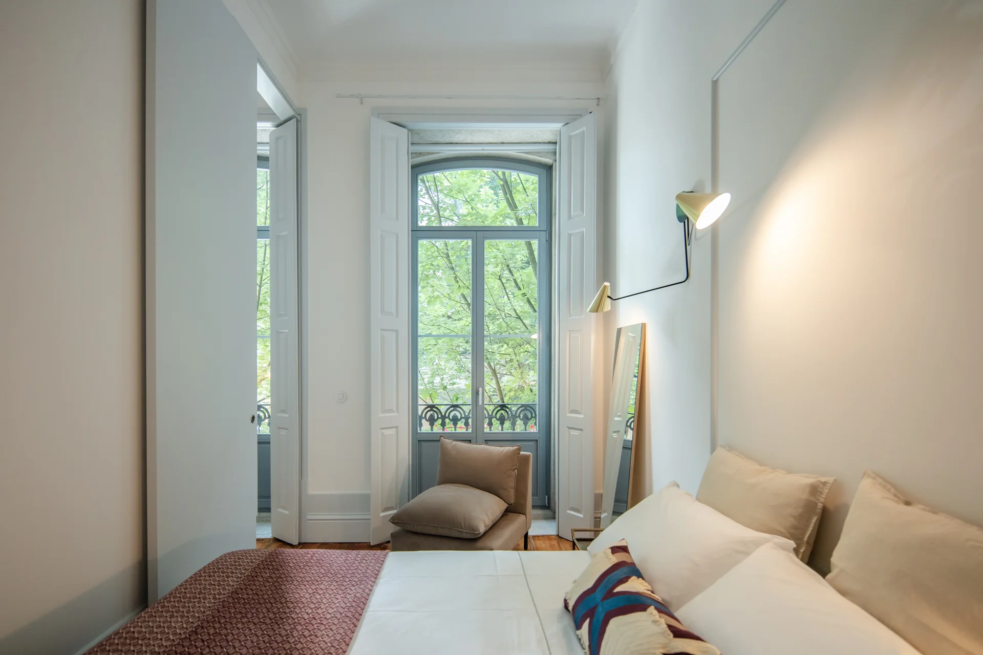 A bright bedroom featuring a bed with white linens, a tan armchair by a tall window with light blue trim, and a gold swing-arm wall sconce.