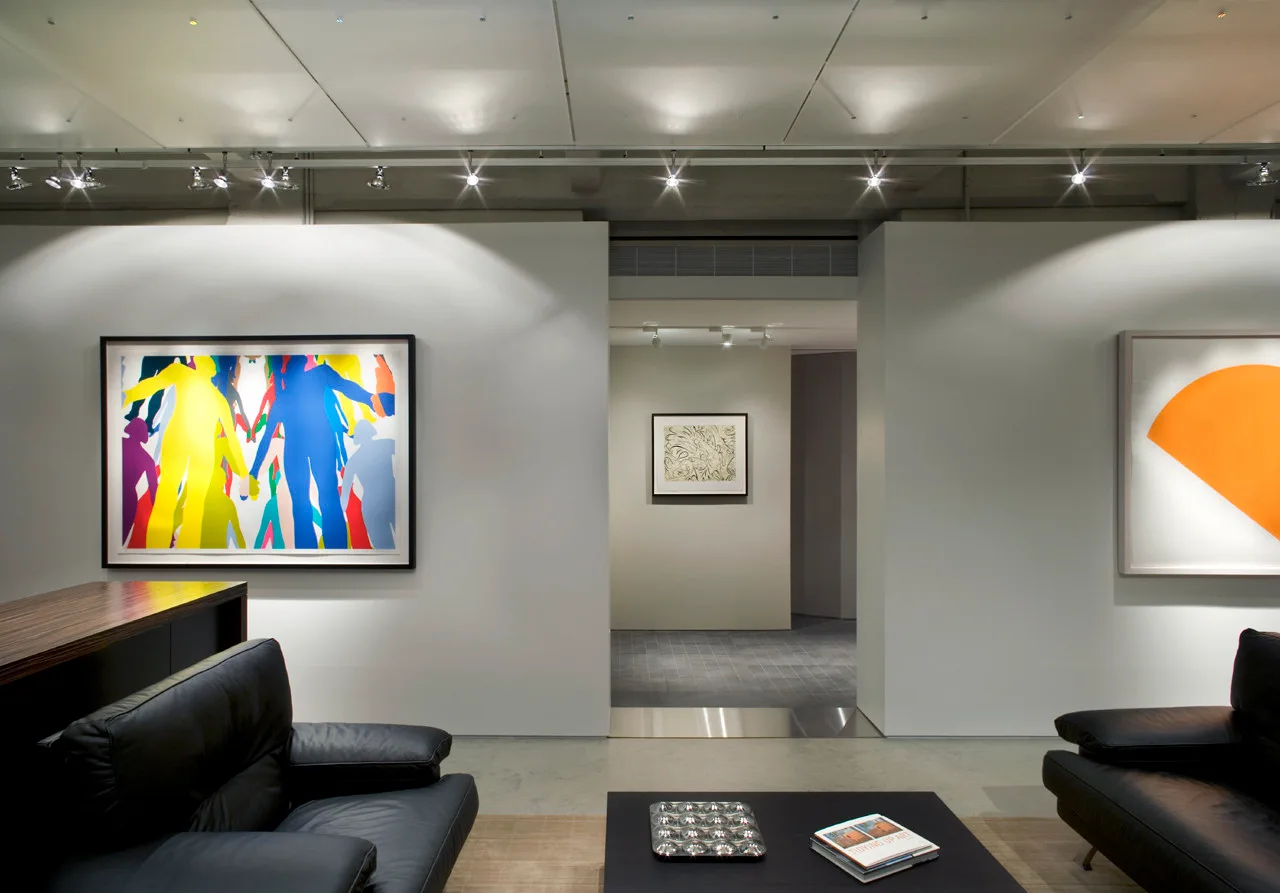A minimalist living room features black leather sofas, a low coffee table, and large colorful framed artwork illuminated by overhead track lighting.