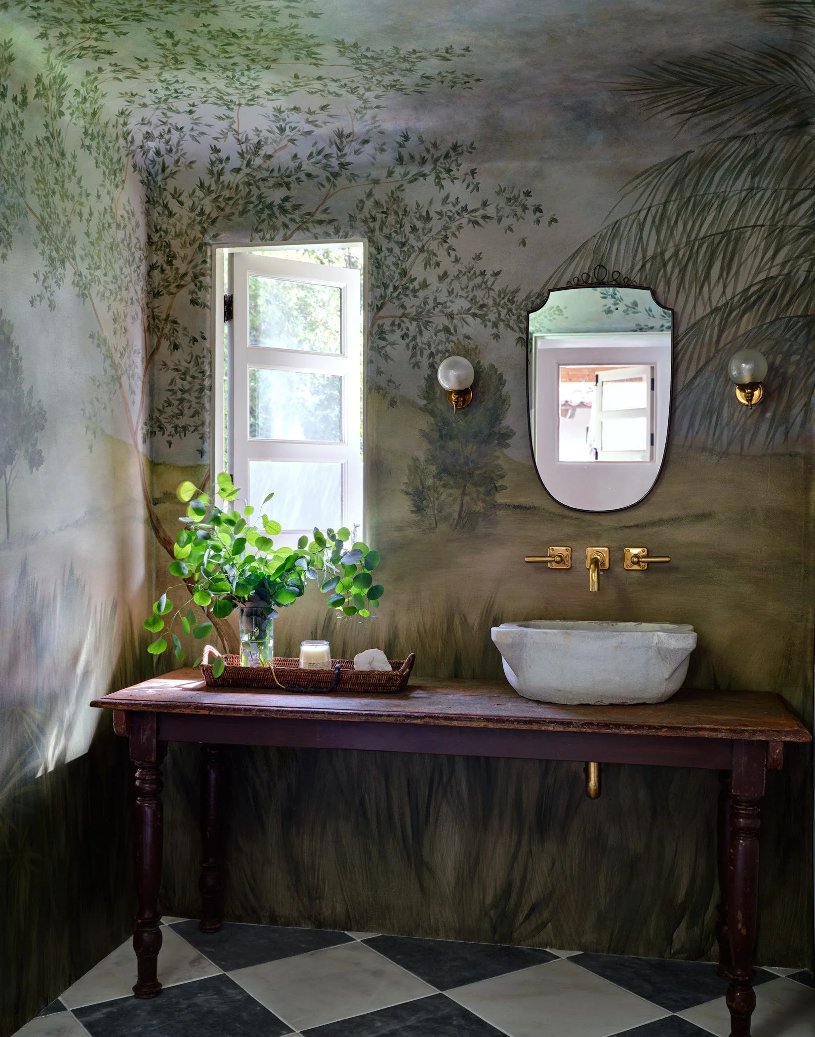 A powder room with landscape mural walls, a rustic wood vanity, a stone vessel sink, brass fixtures, and a black and white checkerboard tile floor.