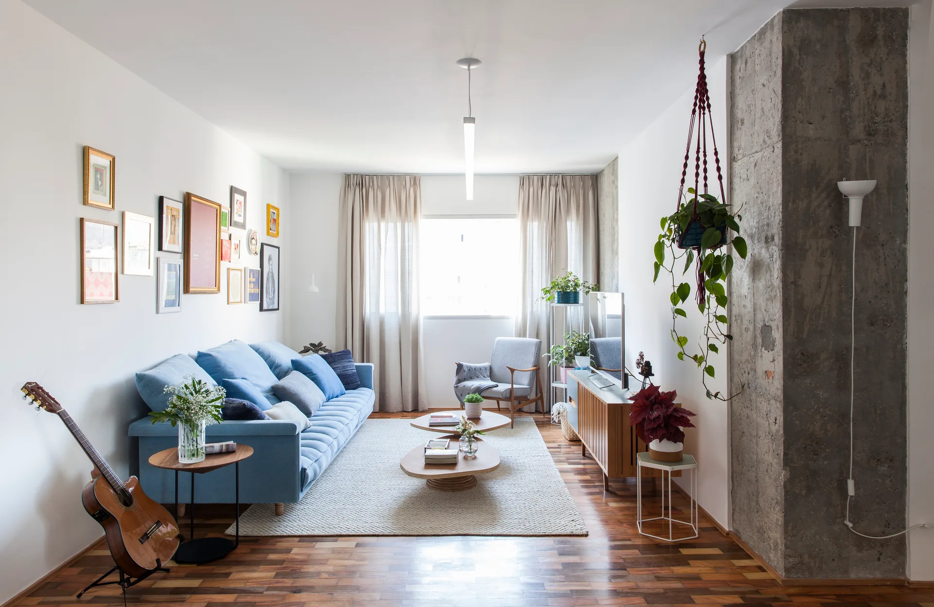 A bright living room features a blue sofa below a gallery wall, curved wood coffee tables, and an exposed concrete pillar next to a wooden console.