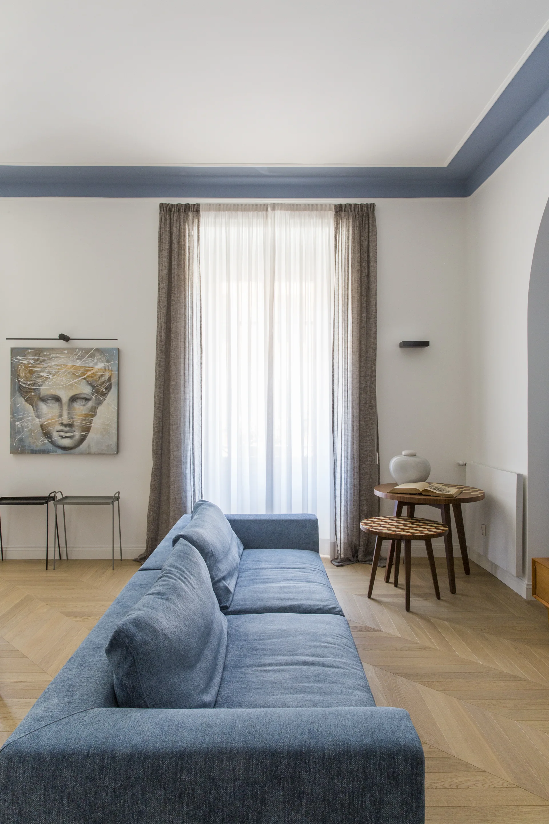 A blue sofa sits on light wood herringbone flooring in a living room with sheer curtains and a blue painted border on the ceiling.