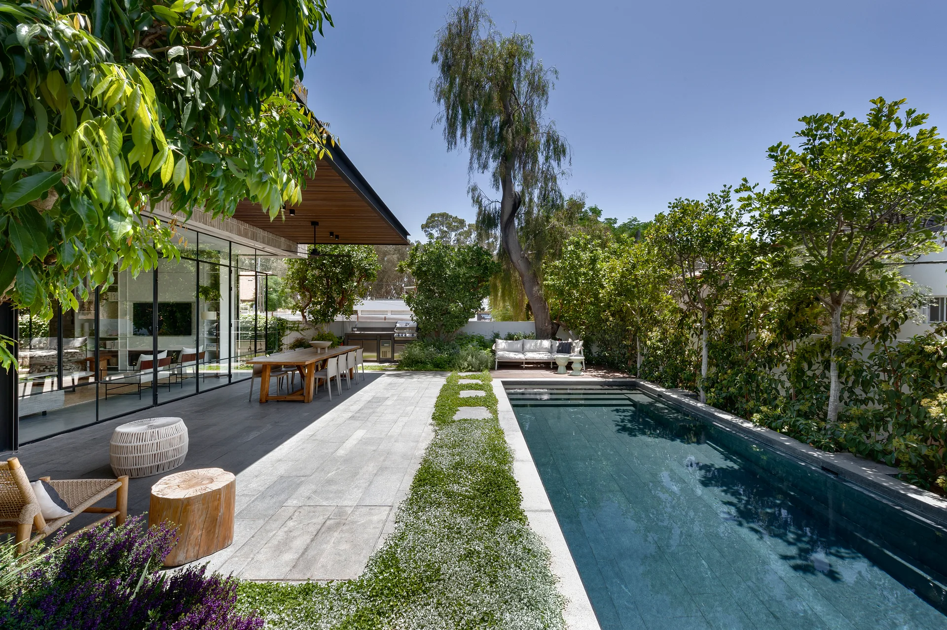A rectangular swimming pool borders a stone tile patio featuring a wood dining table set under a slatted ceiling overhang and glass-walled home.
