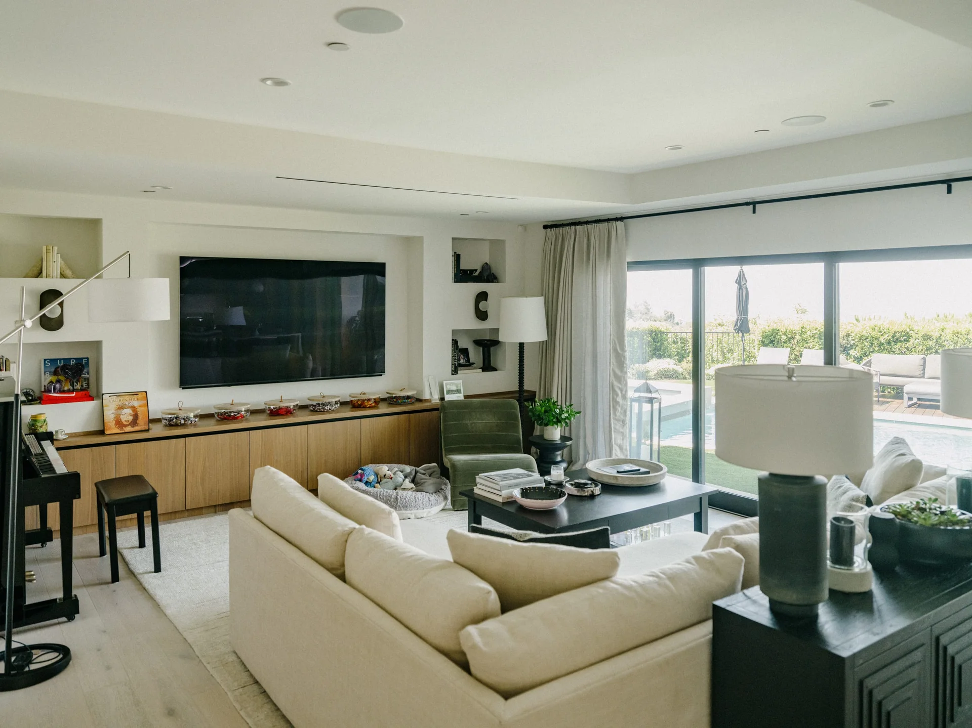 A bright living room features a cream sectional sofa, a black coffee table, a low wooden media console, and large sliding glass doors.