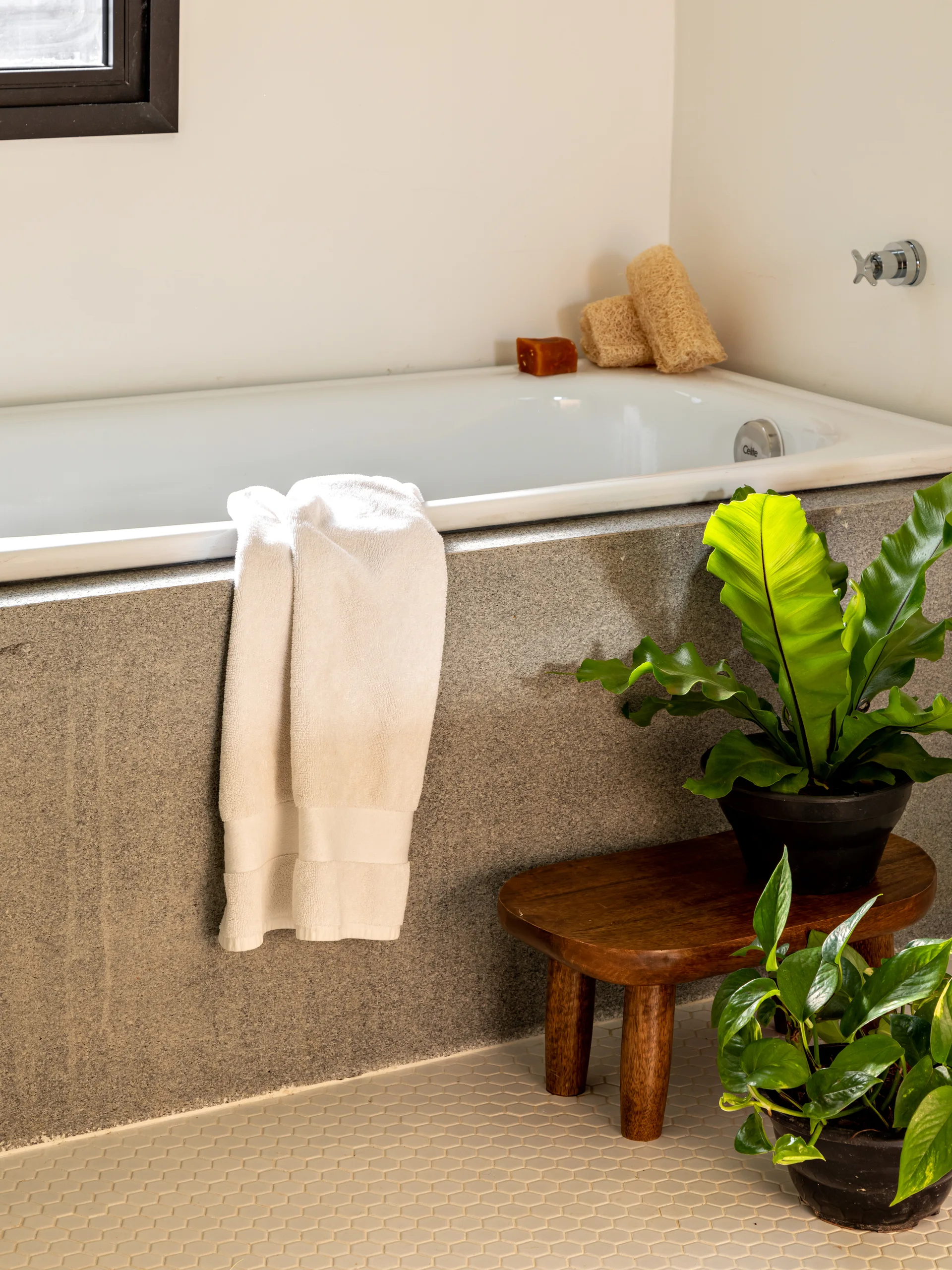 A white built-in bathtub with a grey stone surround features a draped white towel, a low wooden stool with potted plants, and hexagonal floor tiles.