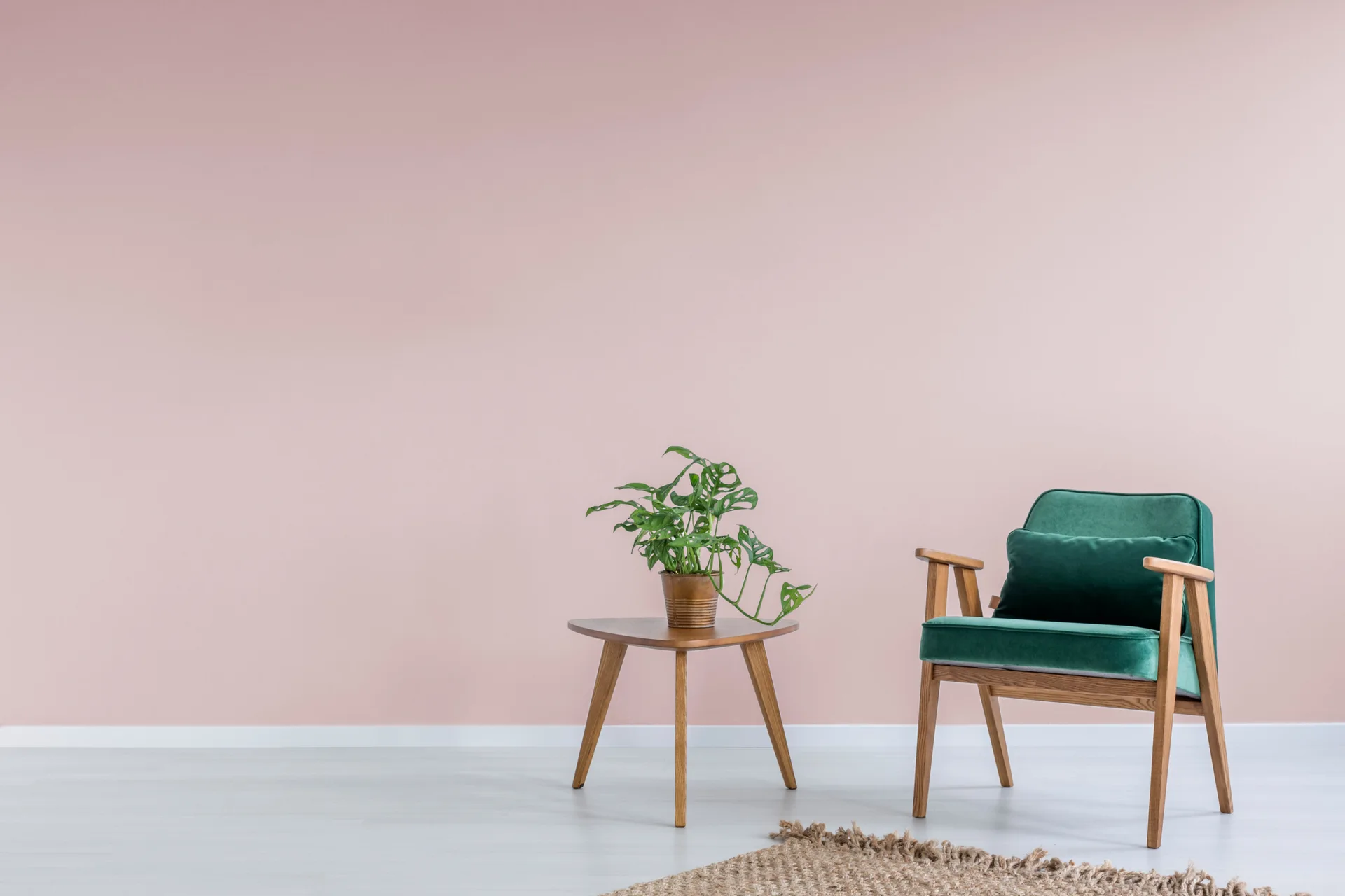 A green velvet armchair stands beside a wooden side table with a potted plant against a matte pink wall above a light floor.
