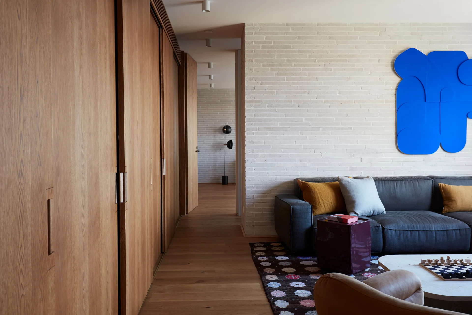 A modern living room with a charcoal gray sofa, a white painted brick wall with large blue artwork, and a full wall of warm wood cabinets.