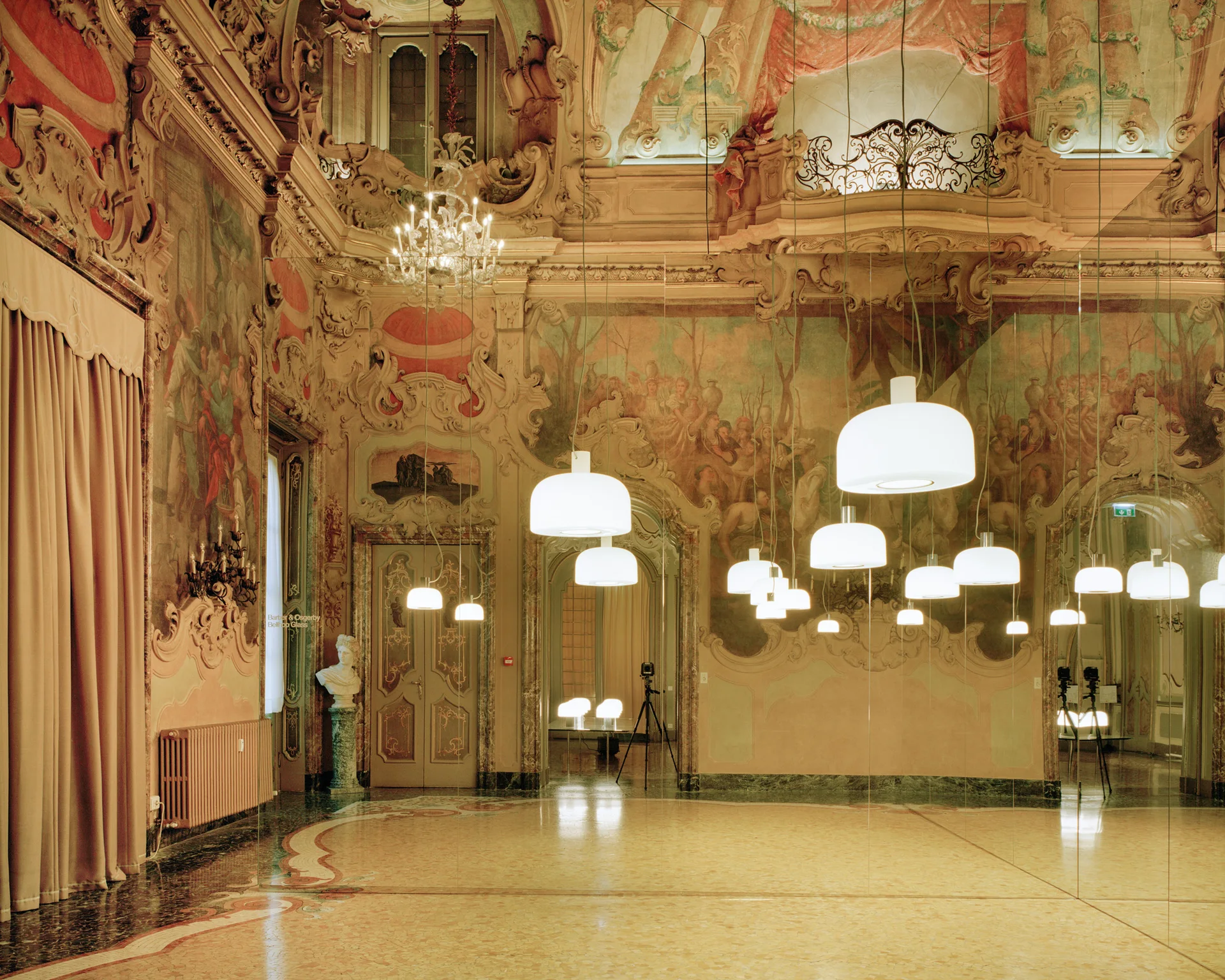 A grand hall featuring ornate wall frescoes, classical moldings, and polished floors is illuminated by multiple modern white dome pendant lights.