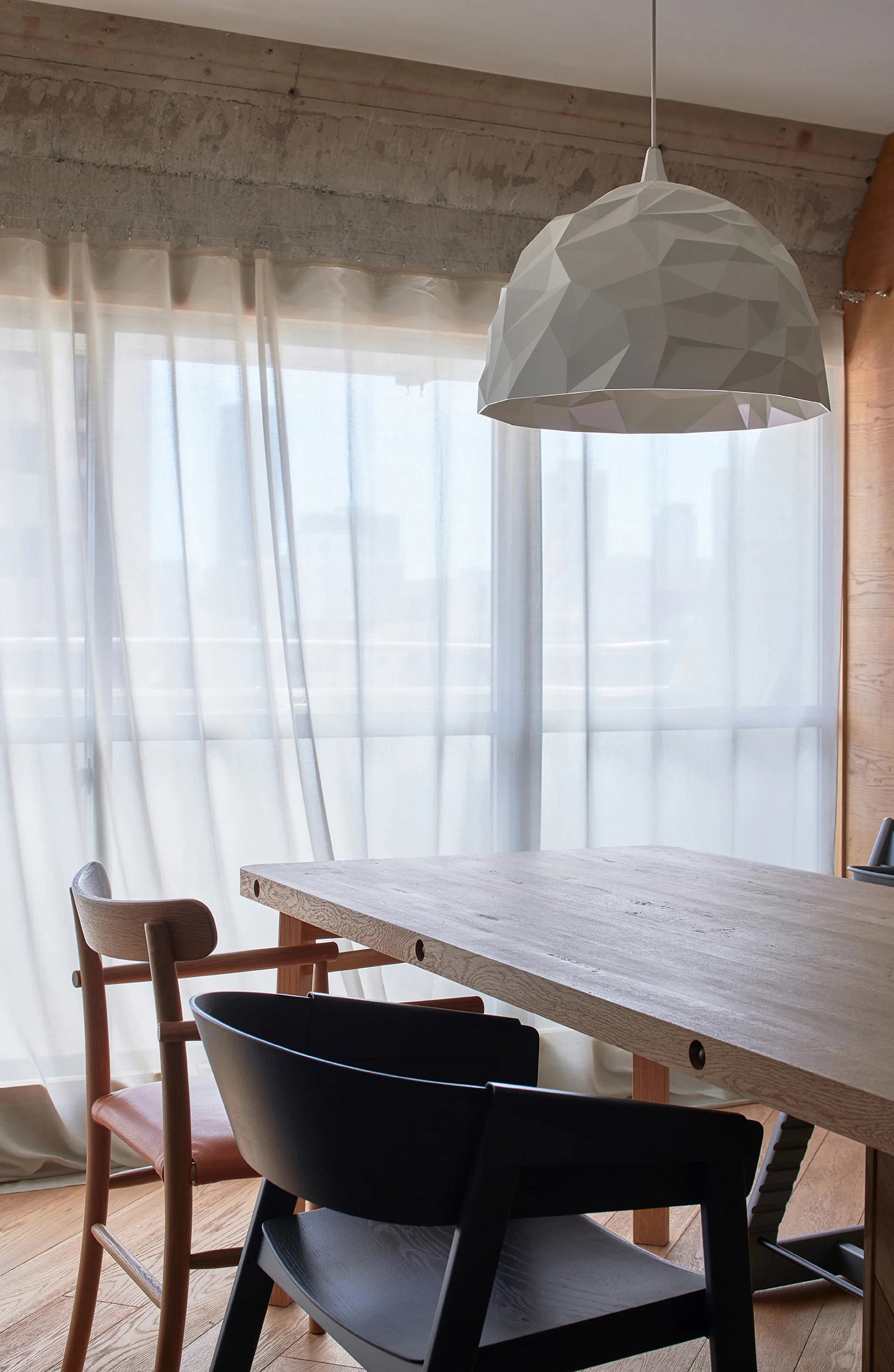 A modern dining room features a wood table, a black chair, a wood chair, and a white geometric dome pendant light in front of sheer white curtains.