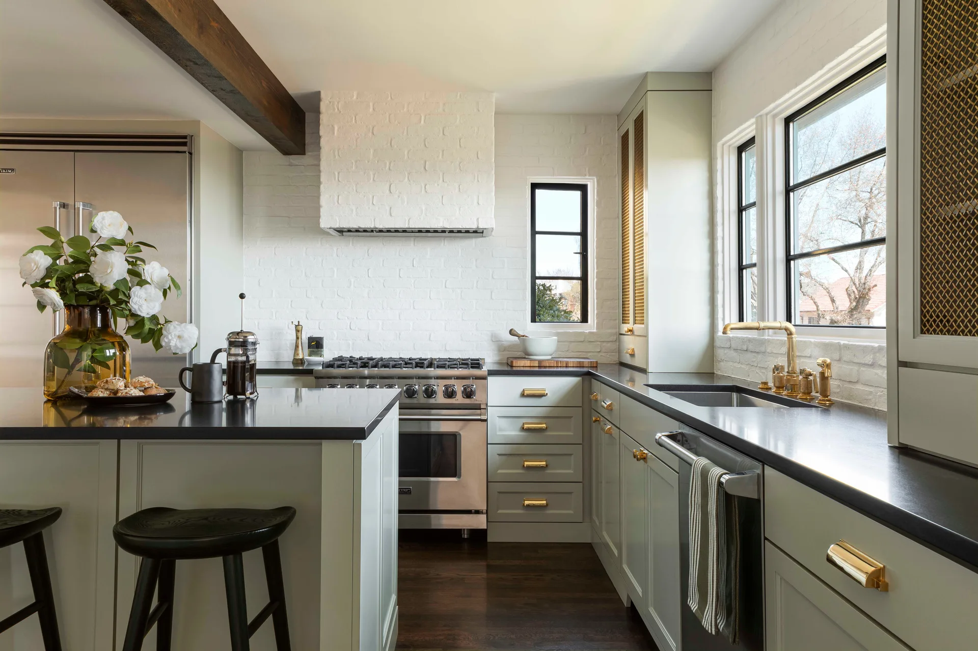 A transitional kitchen featuring sage green cabinets with brass hardware, a white painted brick wall, dark countertops, and a wood ceiling beam.