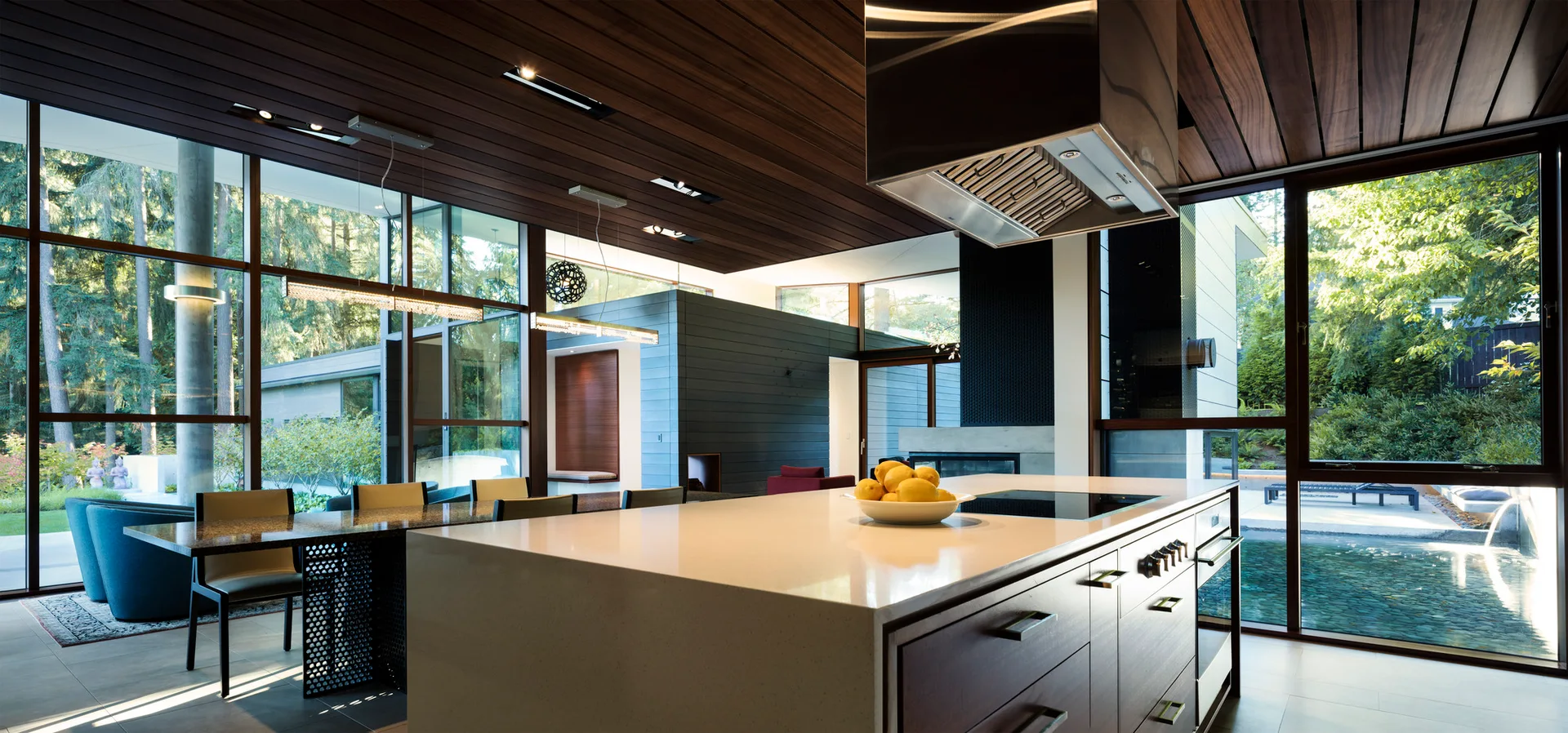 A modern kitchen features a dark wood slatted ceiling, a large white-topped island with a range, and floor-to-ceiling windows showing an outdoor pool.