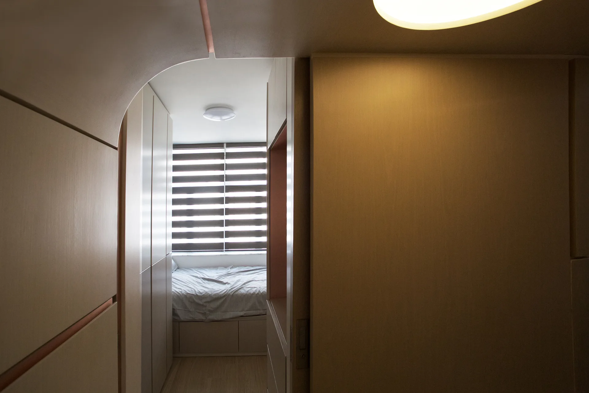 A view through an asymmetrical arched doorway into a compact bedroom with a platform bed, striped blinds, and built-in wood cabinetry.