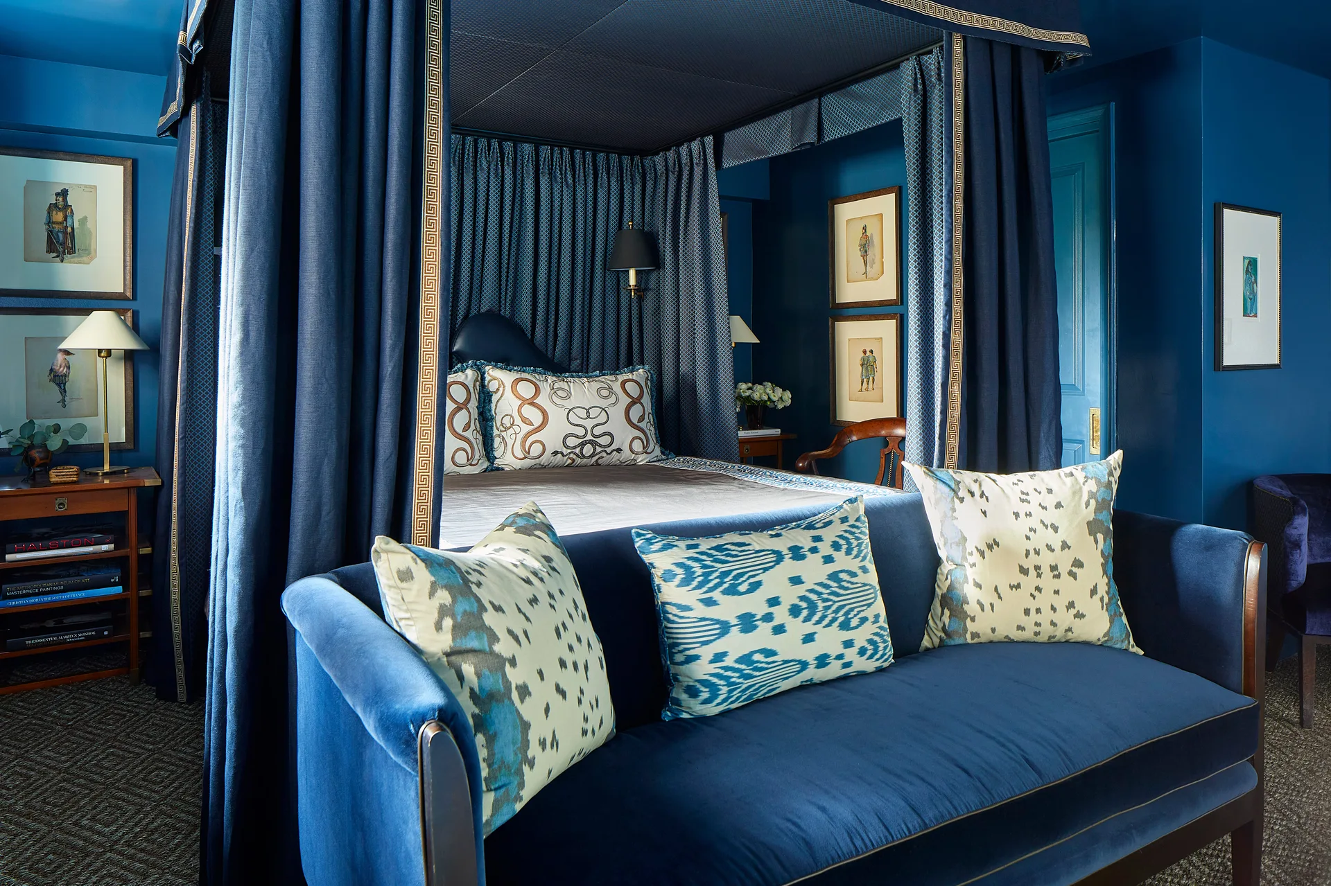 A monochromatic deep blue bedroom features a large canopy bed with heavy blue drapes and a blue velvet sofa with patterned throw pillows in the foreground.