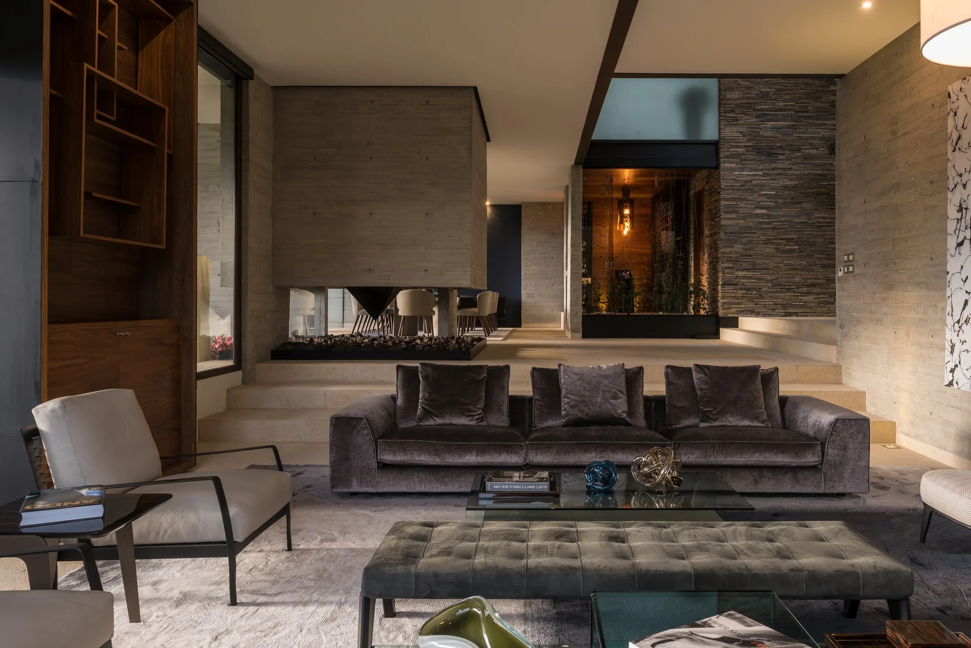 A sunken living room features a dark velvet sofa, a tufted ottoman, raw concrete walls, and stone steps leading to an upper level.