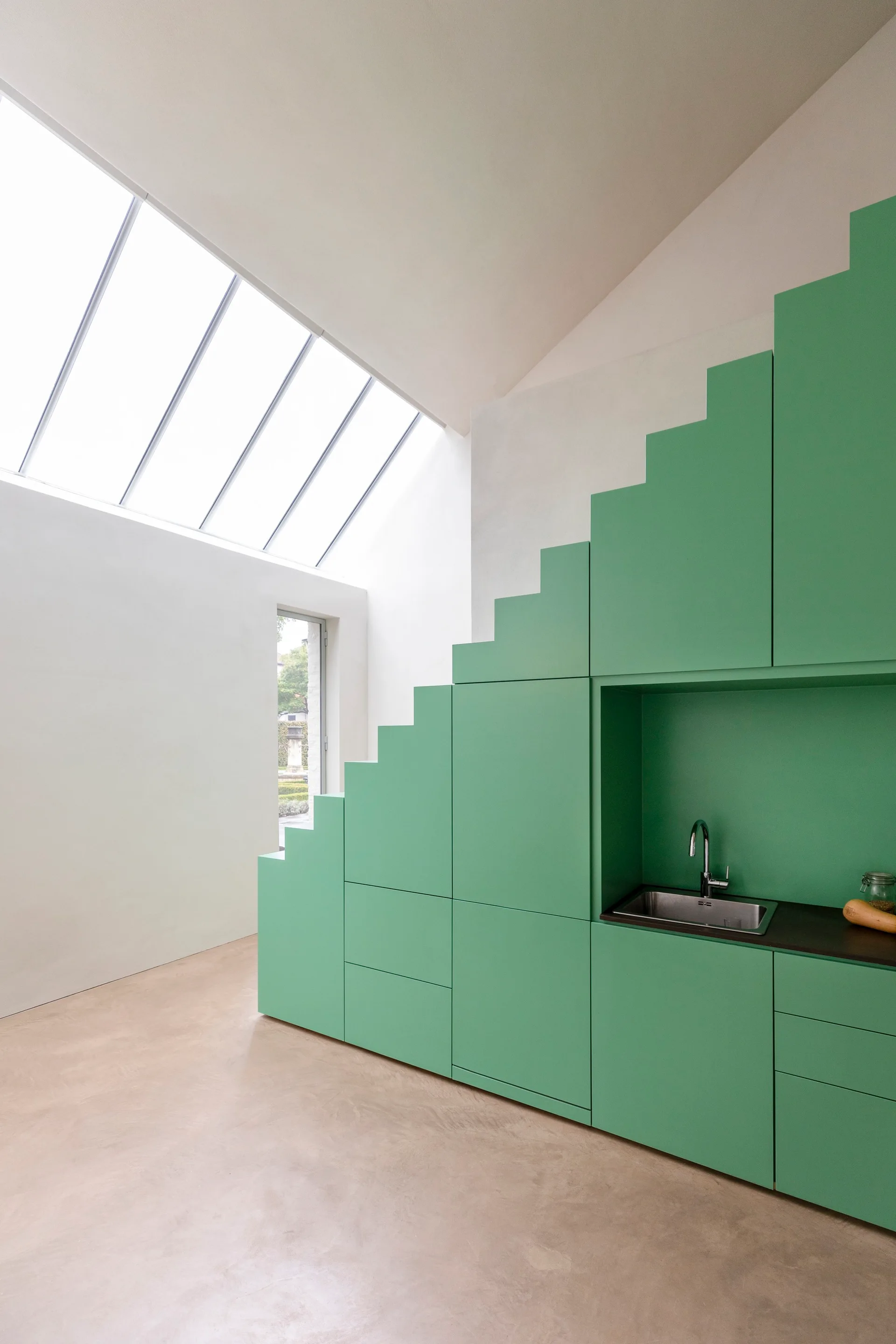A minimalist kitchen features a large stepped mint green cabinet unit, an integrated stainless steel sink, and a slanted skylight on the ceiling.