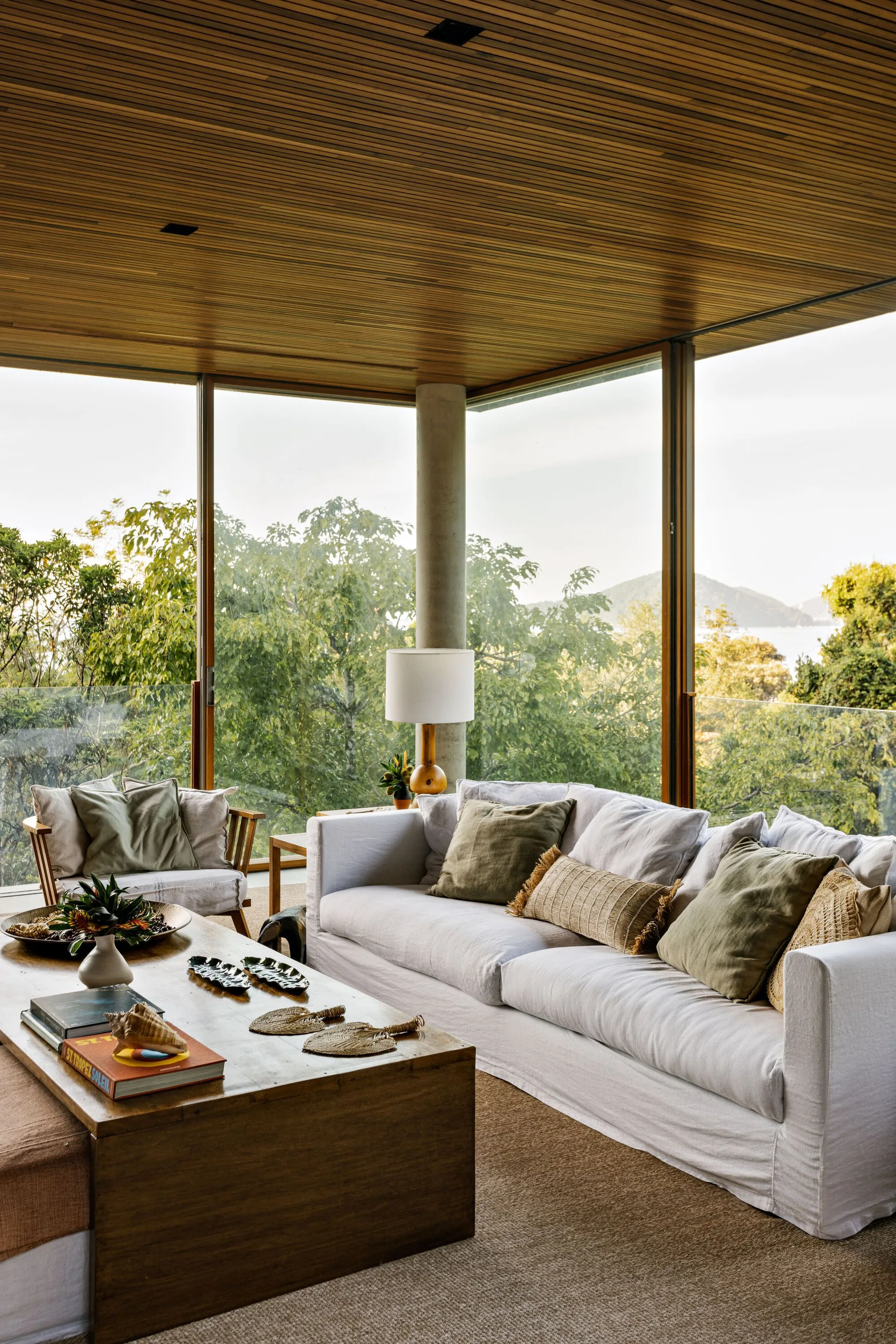 A modern living room features a white linen sofa, a low wooden coffee table, and glass corner walls under a slatted wood ceiling.
