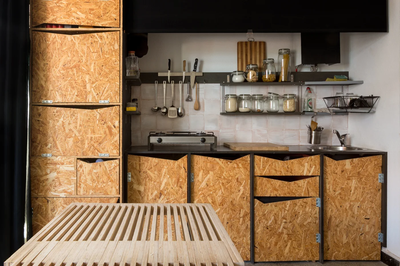 A small kitchen features raw oriented strand board cabinets, a white tiled backsplash, open metal shelves, and a slatted wood table.