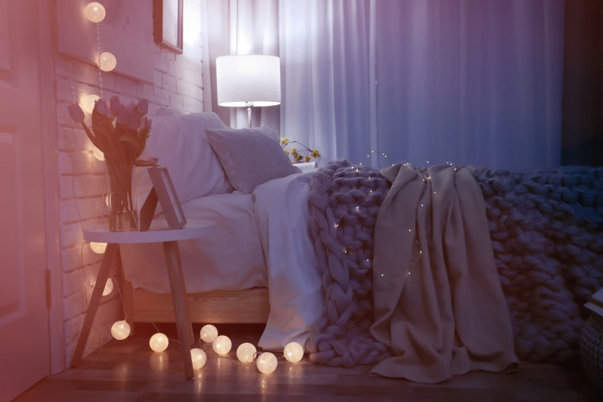A bed with a heavy grey knit blanket and white pillows sits next to a round nightstand with flowers and glowing globe string lights.