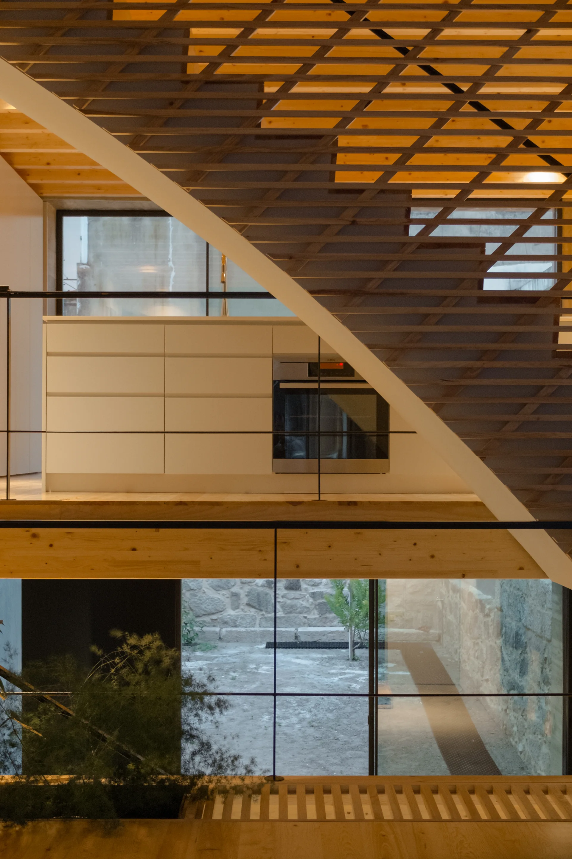 A wooden lattice staircase climbs past a minimalist white kitchen cabinet unit, overlooking a lower level window with a view of a stone courtyard.