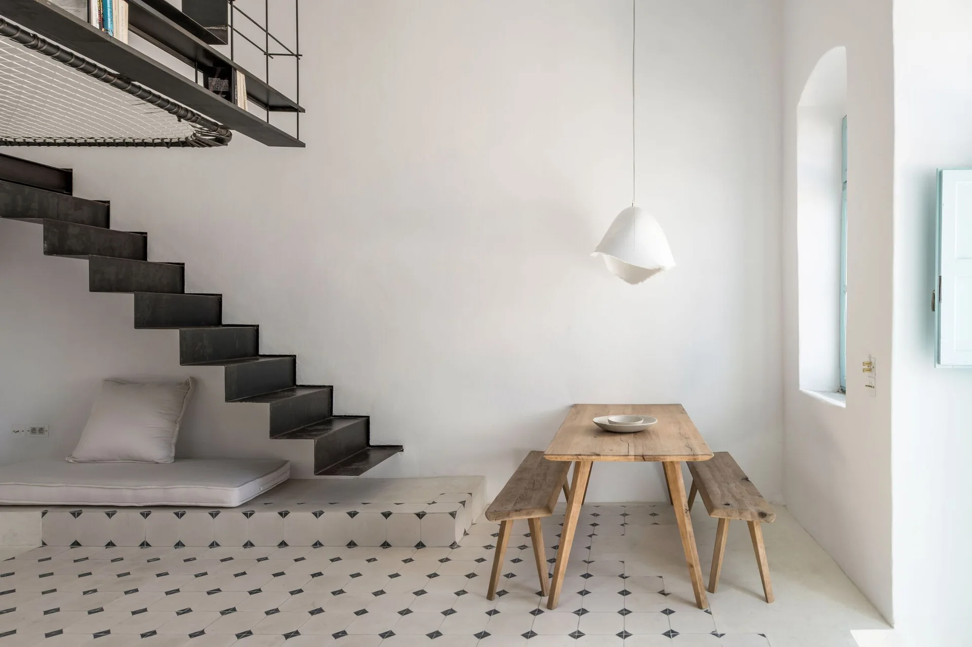 A minimalist dining room features a rustic wooden table and benches beside a black metal floating staircase with a lounge cushion underneath.