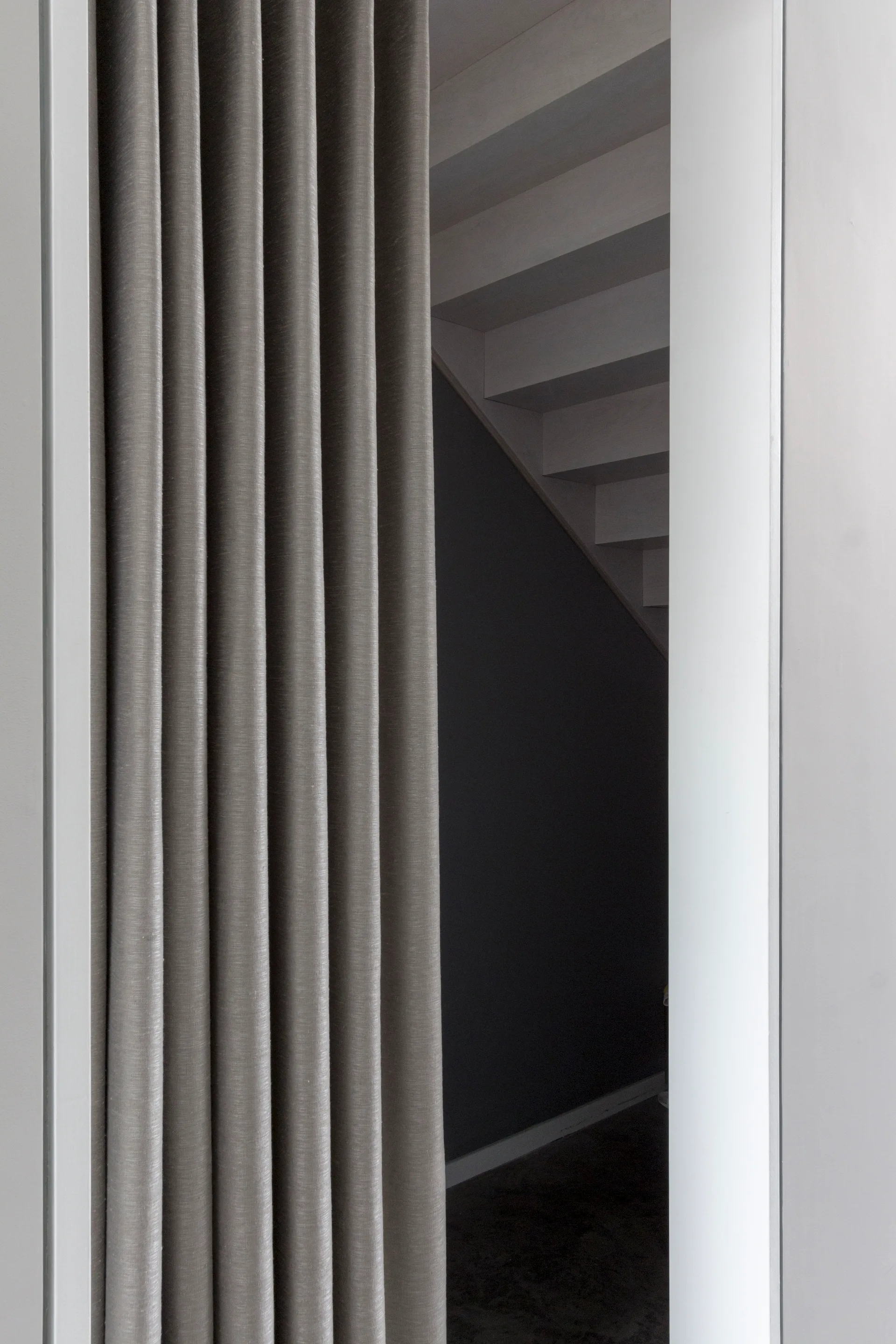 A tall grey pleated curtain hangs in a white doorway, partially obscuring a minimalist staircase in the background.