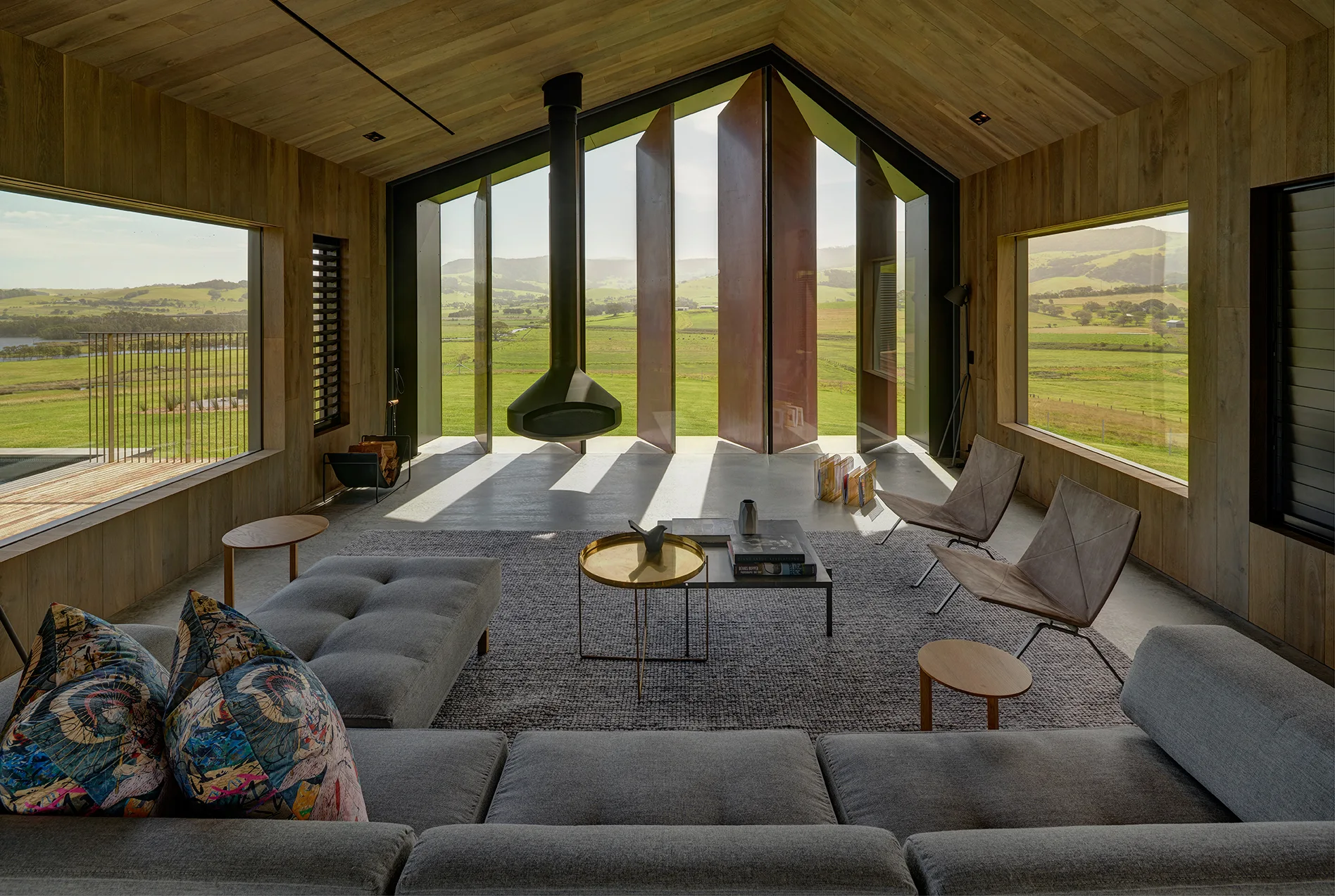 A modern wood-clad living room features a suspended black fireplace, a grey sectional, and pivoting doors facing rolling green hills.