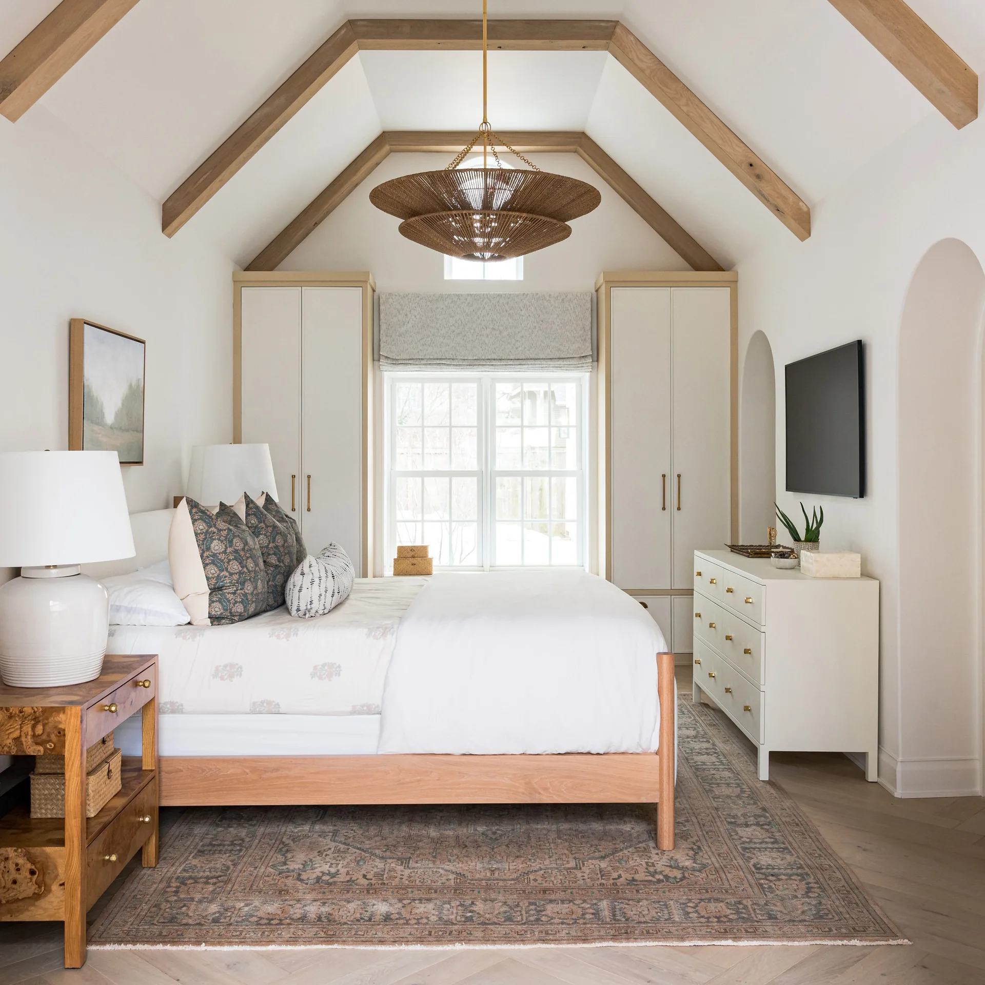 A bedroom features a vaulted ceiling with wood beams, a central bed, and matching white wardrobes framing a window.