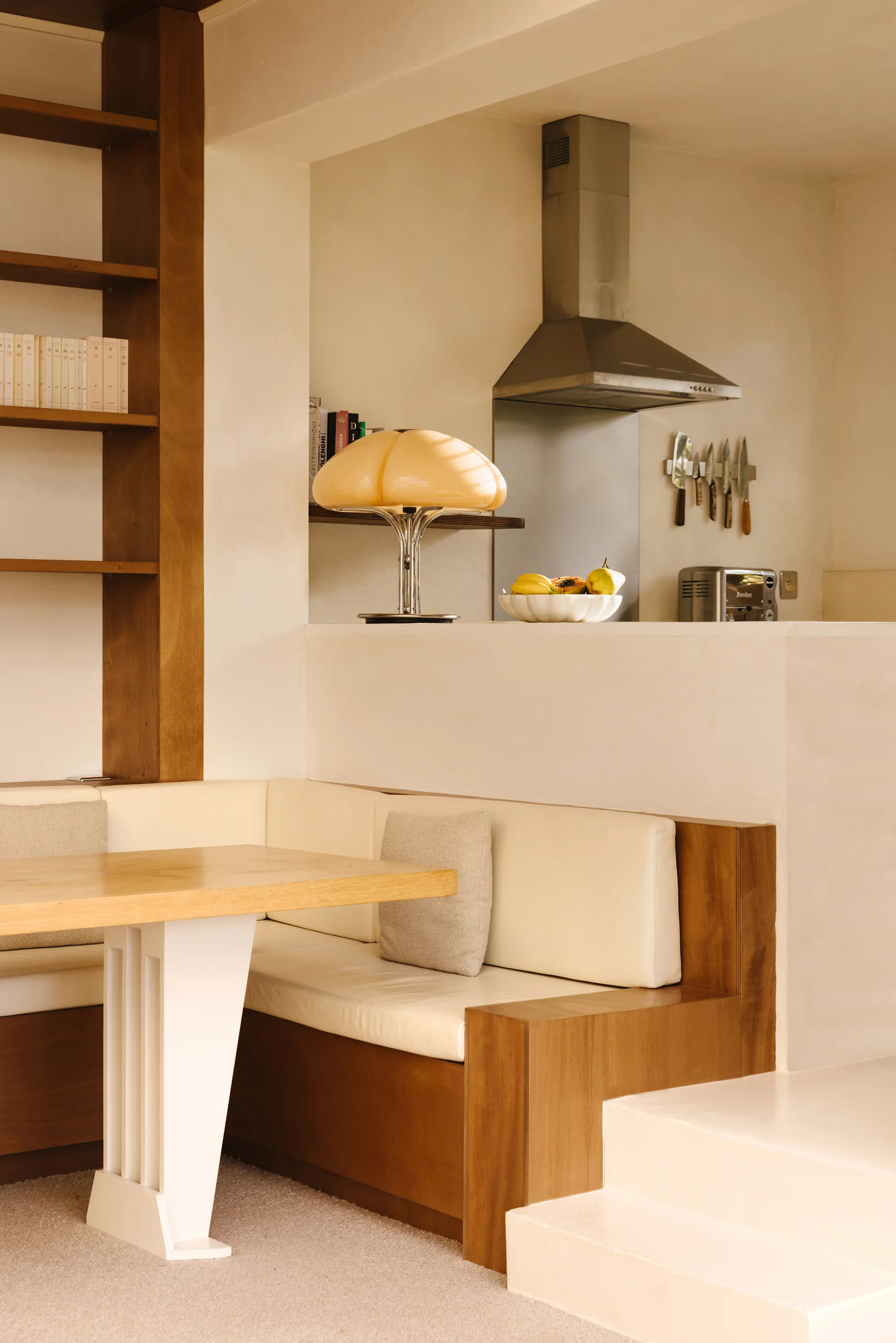 A dining nook with a built-in wood bench and cream cushions sits below a raised kitchen featuring a stainless steel range hood and a mushroom lamp.