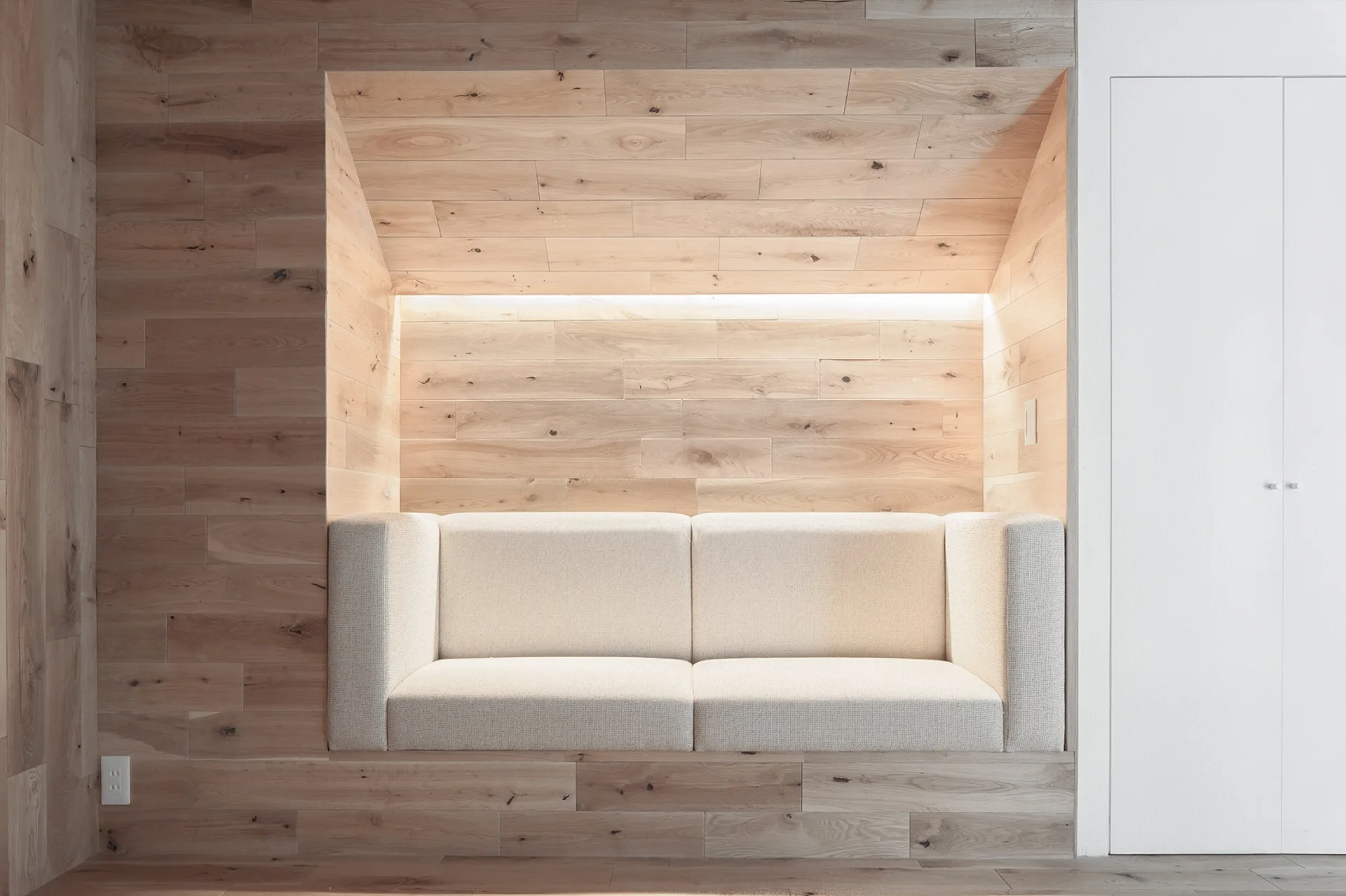 A beige fabric sofa is built into a recessed niche lined with horizontal light wood paneling and illuminated by warm cove lighting.