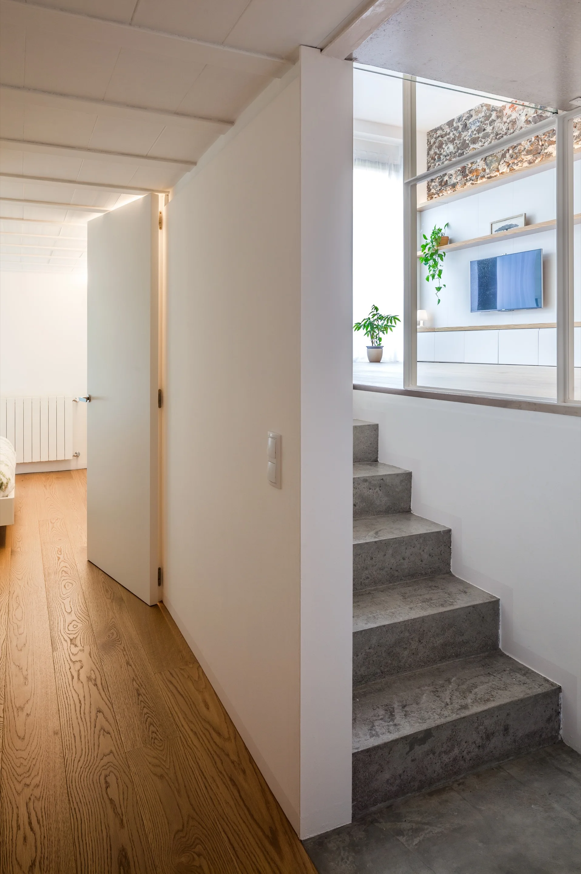A minimalist hallway with light wood flooring leads to concrete stairs, with a glass partition showing an upper-level living space.