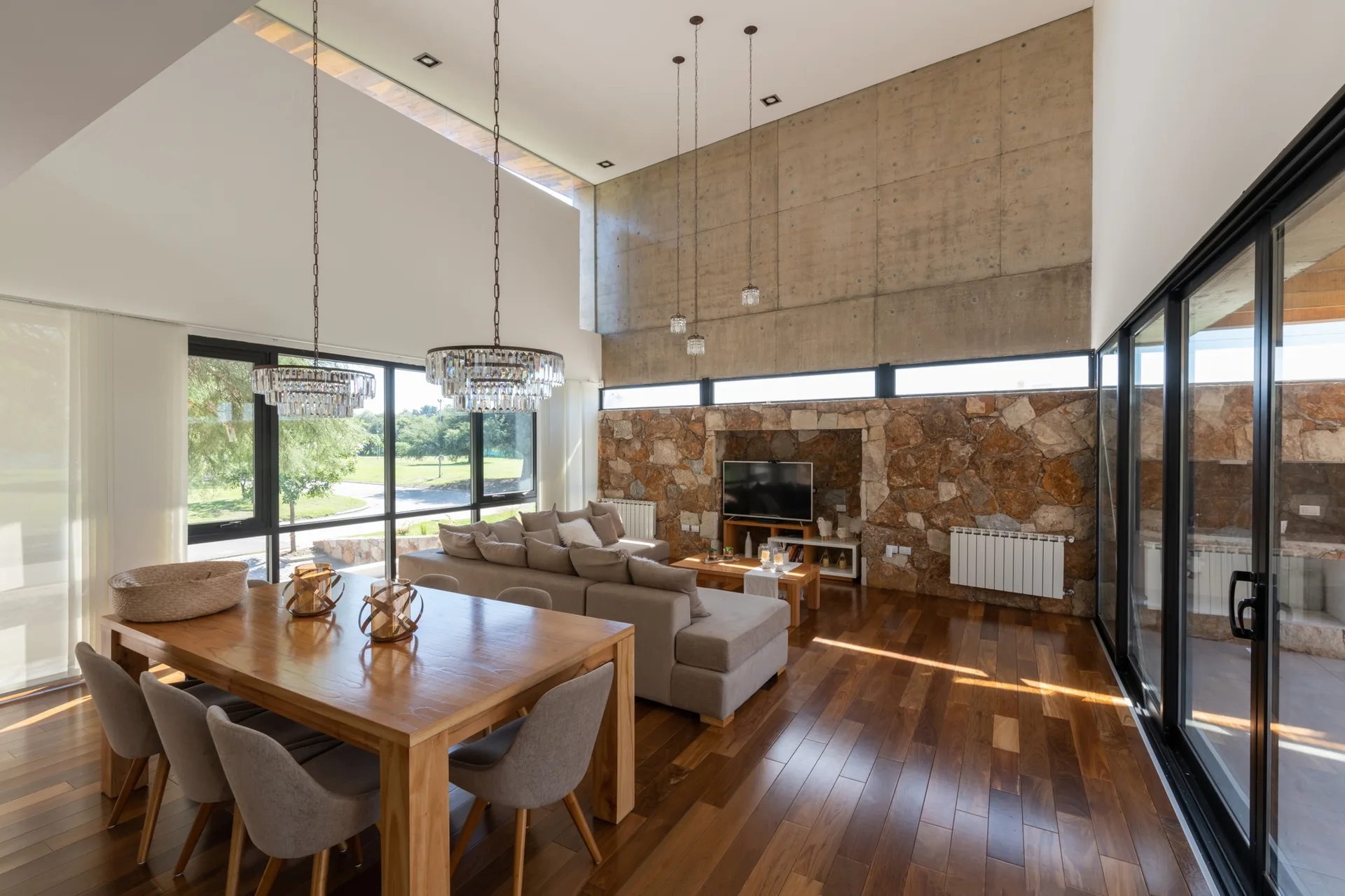 An open-plan living and dining room featuring a high concrete and stone wall, a large wooden table, a grey sectional sofa, and glass sliding doors.