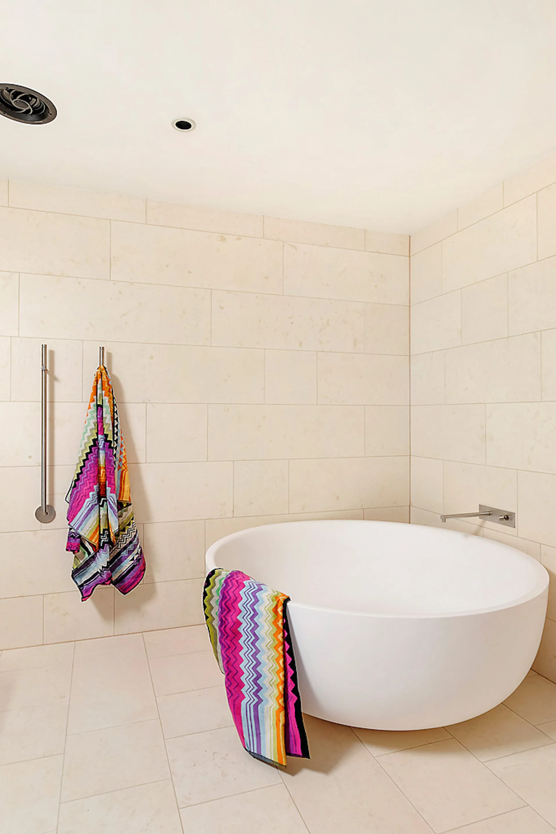 A minimalist bathroom with a round white freestanding bathtub, beige tiled walls, and colorful chevron pattern towels.