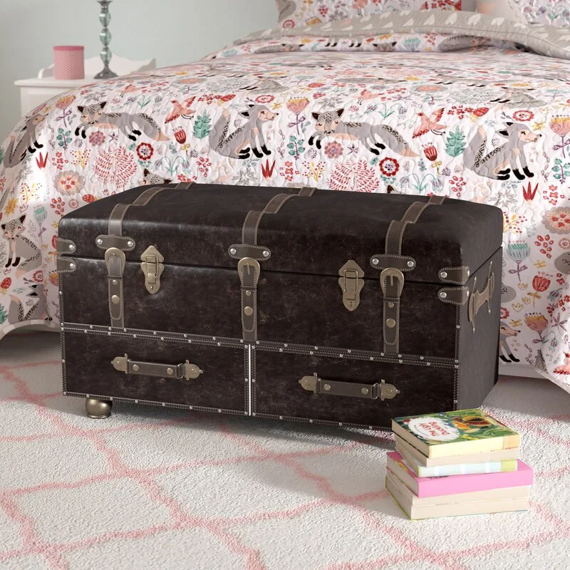 A dark brown faux-leather storage trunk with brass latches sits at the foot of a bed covered in a patterned fox quilt on a pink and white rug.