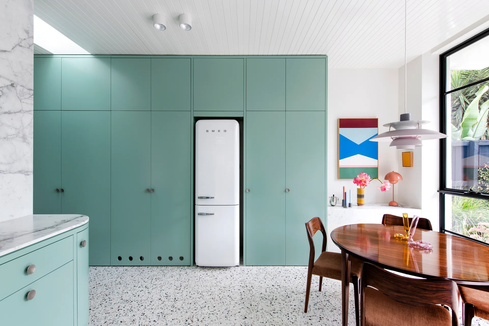 A modern kitchen dining area features a wall of mint green built-in cabinets, an integrated white refrigerator, terrazzo flooring, and a wood table.