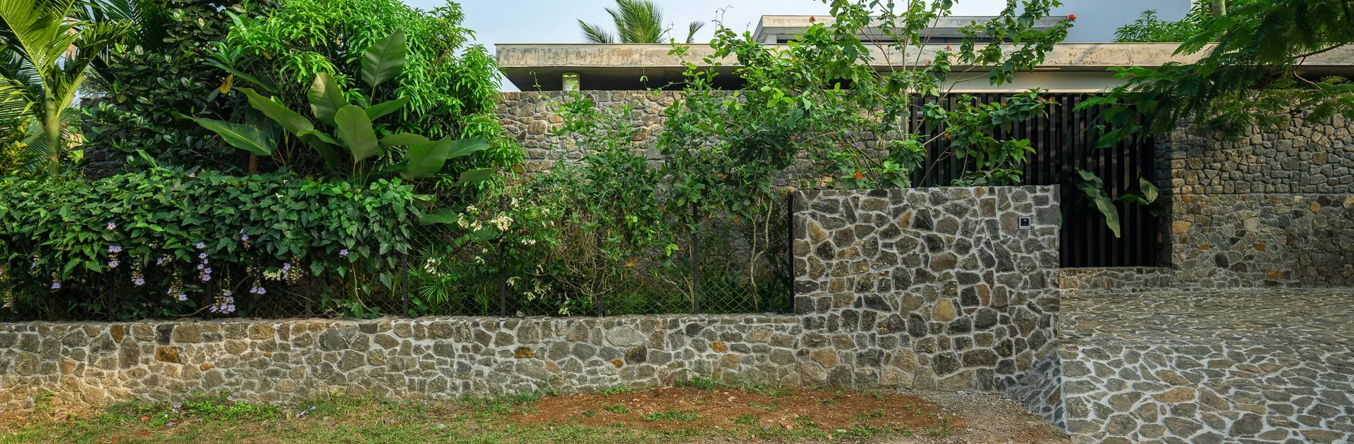 A modern home exterior featuring an irregular stone masonry wall, a matching stone paved driveway, and lush green tropical foliage.