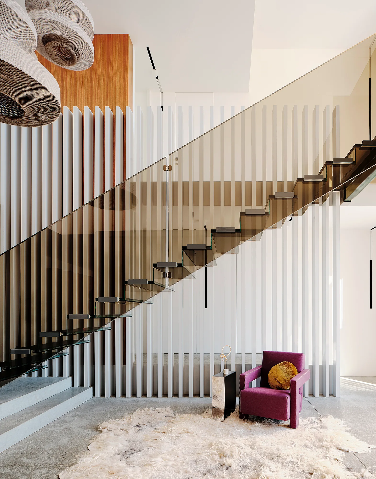 A floating staircase with a tinted glass railing sits in front of a white slatted wall next to a purple armchair and shaggy white rug.