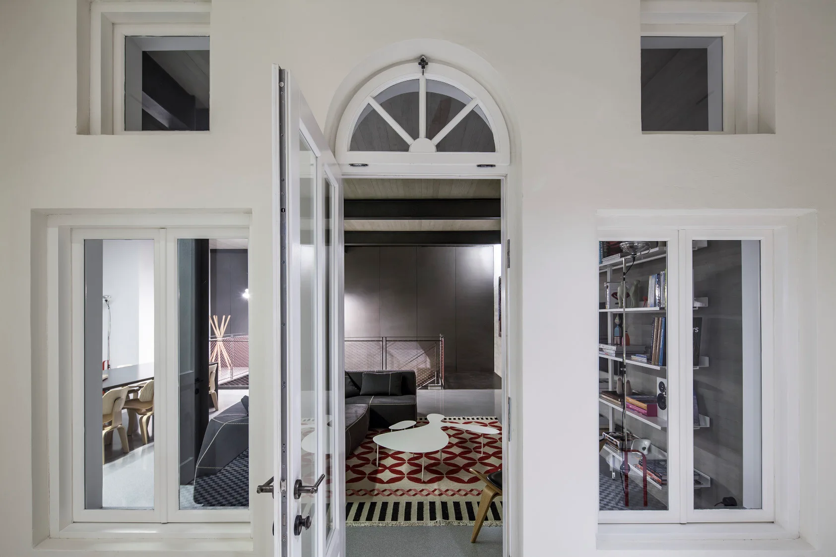 An open arched white door looks into a modern living room featuring a dark grey sectional sofa, a red patterned rug, and irregular white coffee tables.