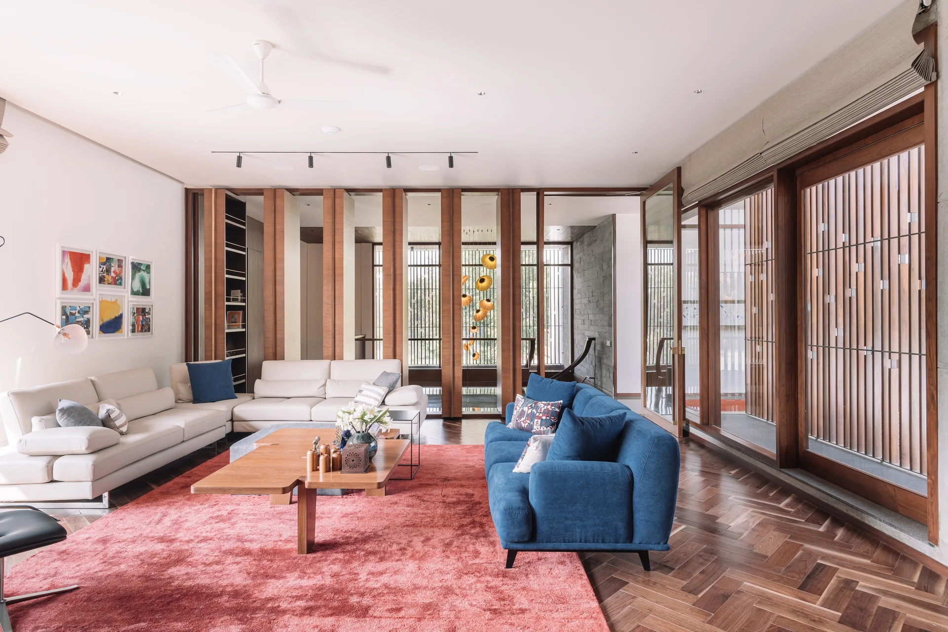 A contemporary living room features a blue sofa, a cream sectional, a red rug on herringbone wood floors, and vertical slatted wood partitions.
